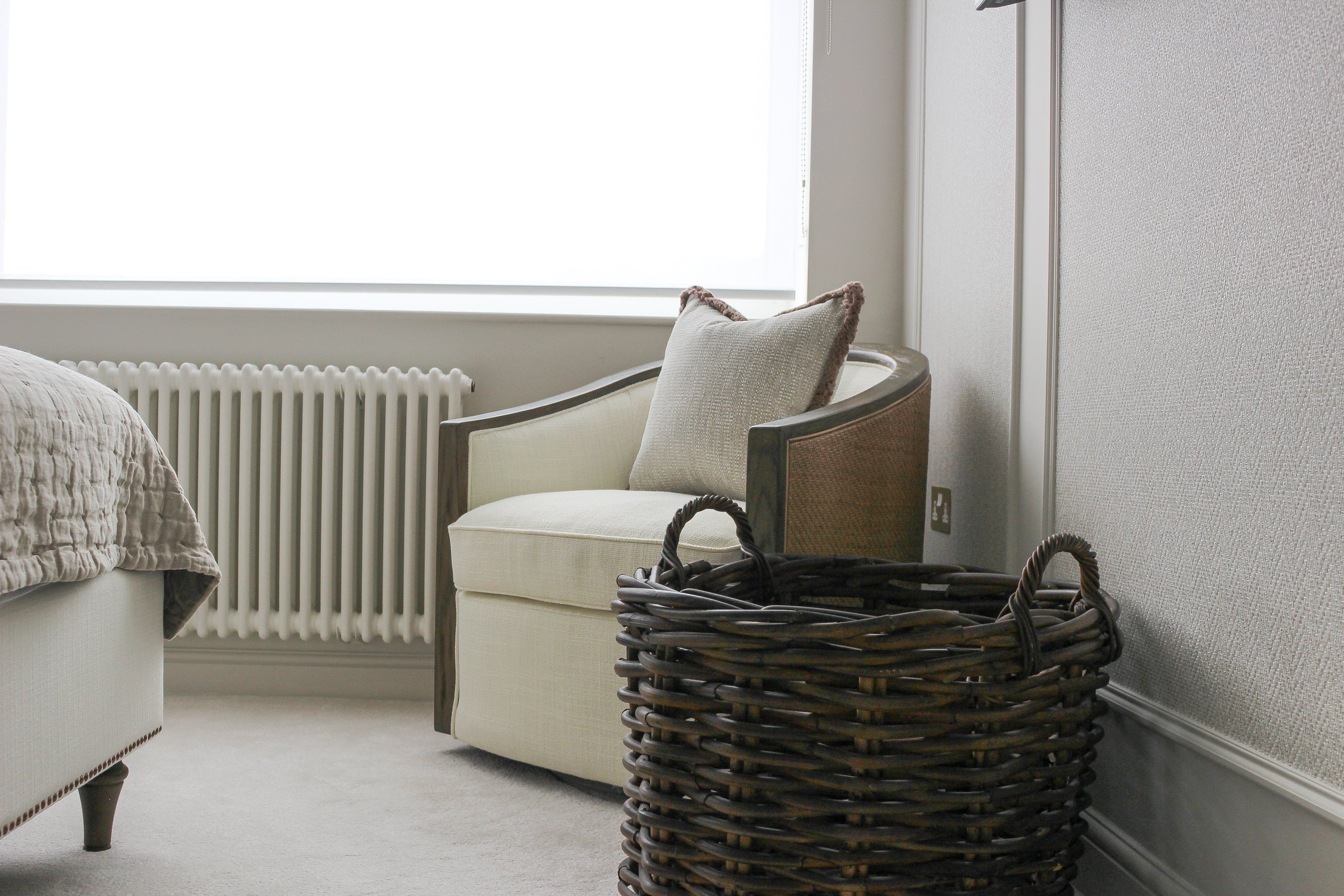 A cream coloured armchair with a textured beige cushion with fringe edging next to a large window, with a large brown wicker basket on the floor nearby. Room designed by Yazzmin Lovelle Interiors 