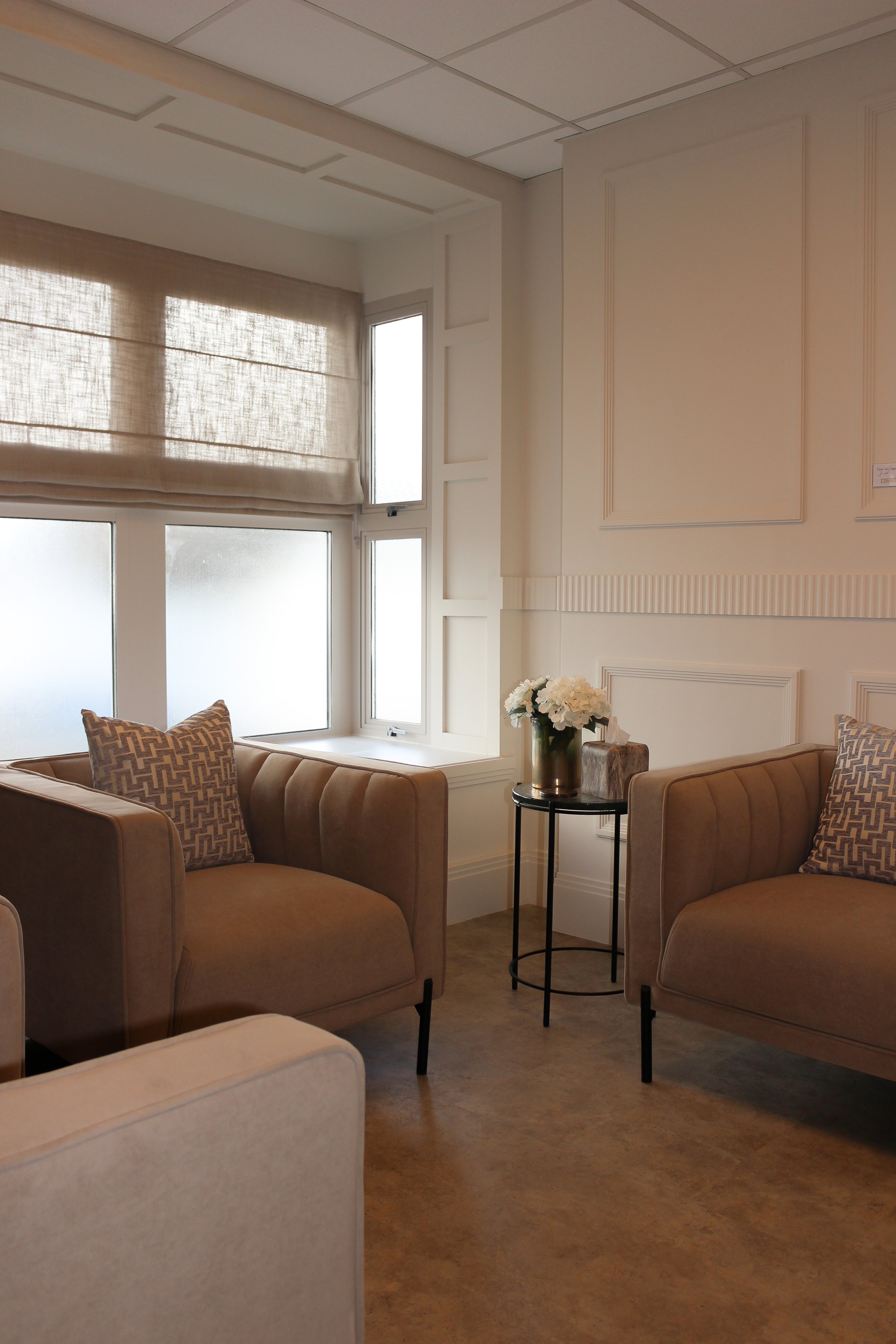 A clinic consultation area with beige armchairs, a side table with white flowers, and frosted windows letting in natural light. Clinic designed by Yazzmin Lovelle Interiors