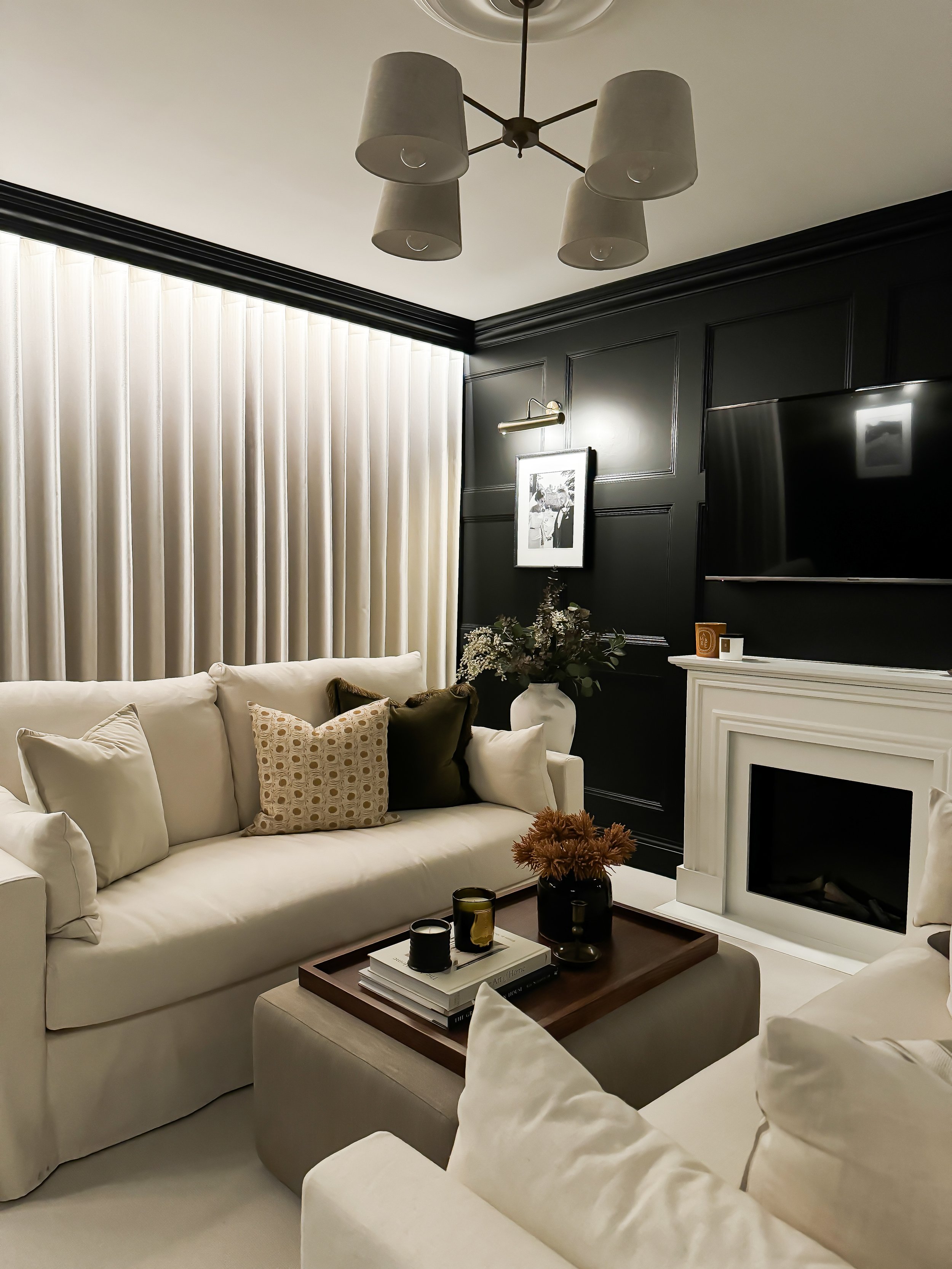 Formal living room with white sofas, a beige ottoman, black wall paneling and a fireplace with a white mantel, decorated with candles, a tray, and flowers. Living room designed by Yazzmin Lovelle Interiors.
