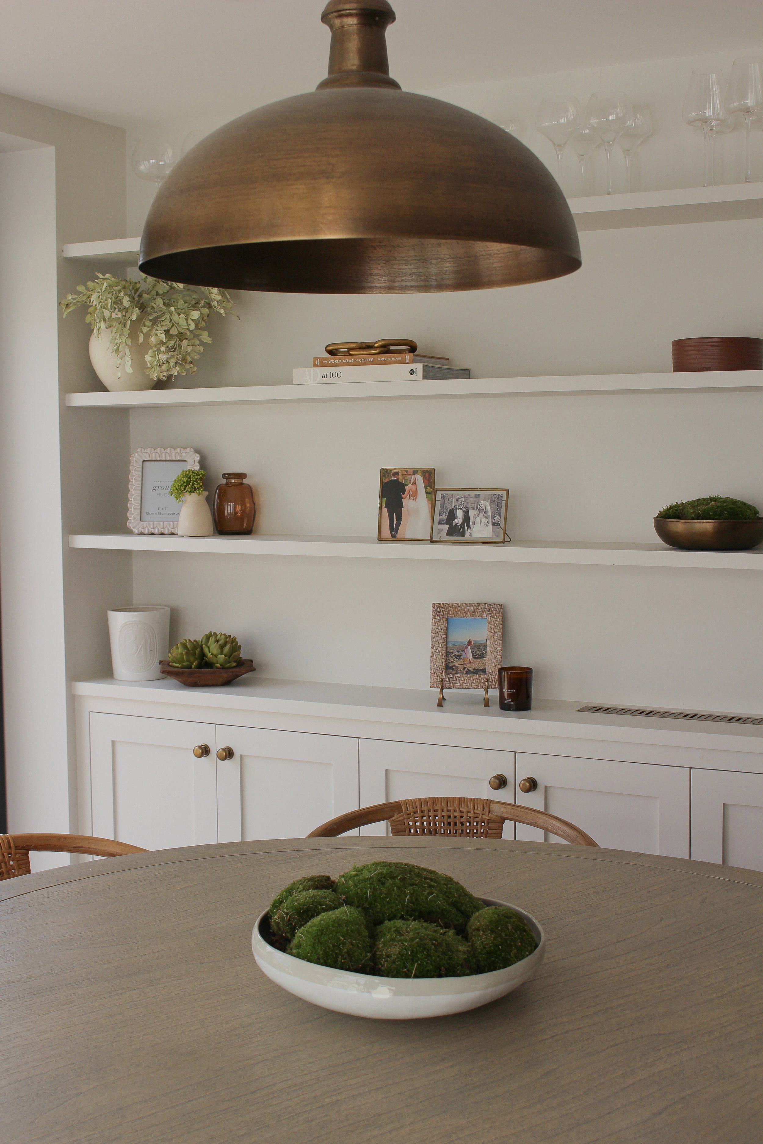 Dining room with a round wooden table, a bowl of moss, a white cabinet with decorative objects, framed photographs, vases, and a large brass pendant light hanging above. Dining area designed by Yazzmin Lovelle Interiors.