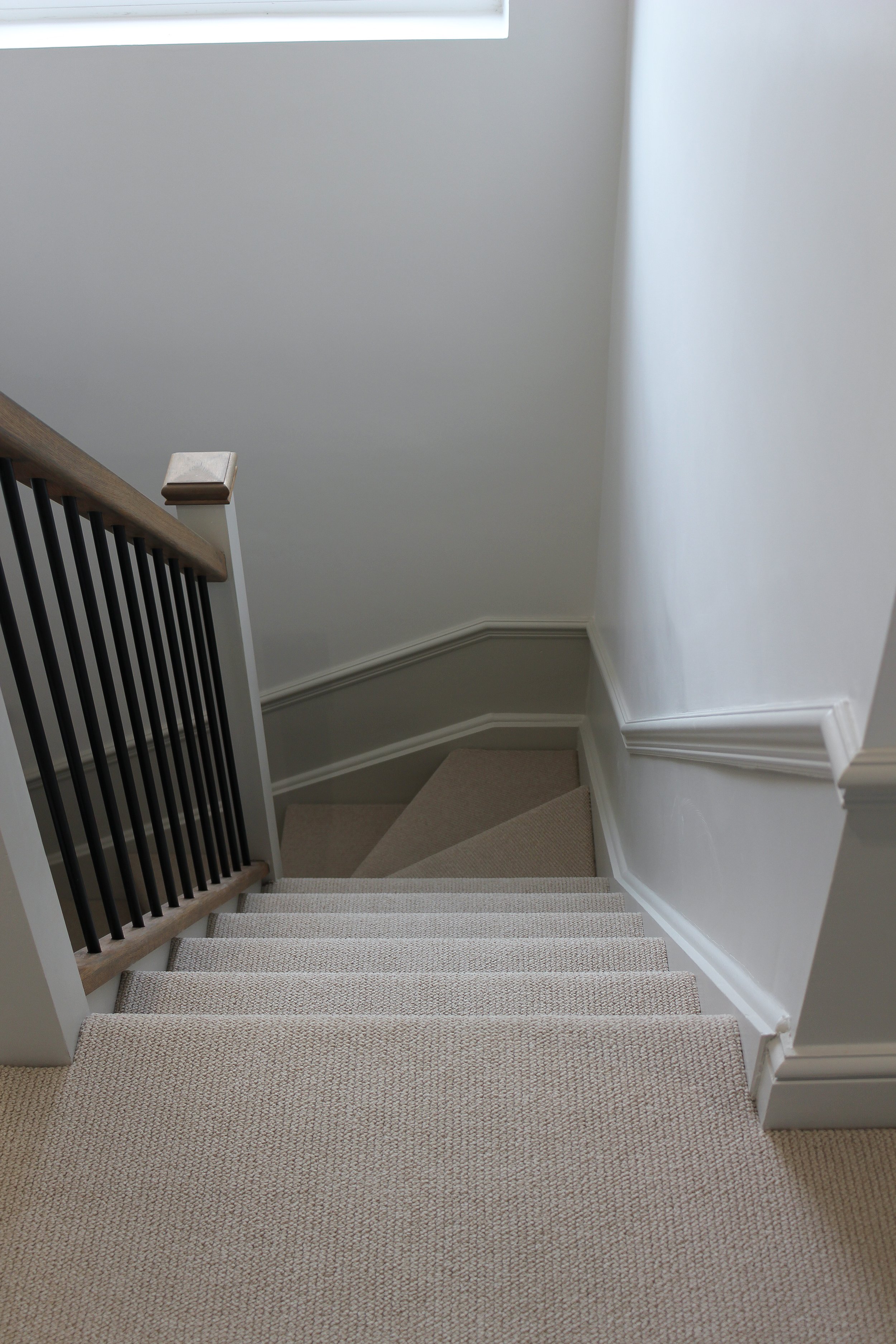 View of a staircase from the top, showing beige carpeted stairs, a wooden handrail with black spindles, white walls, and a window at the top of the staircase.