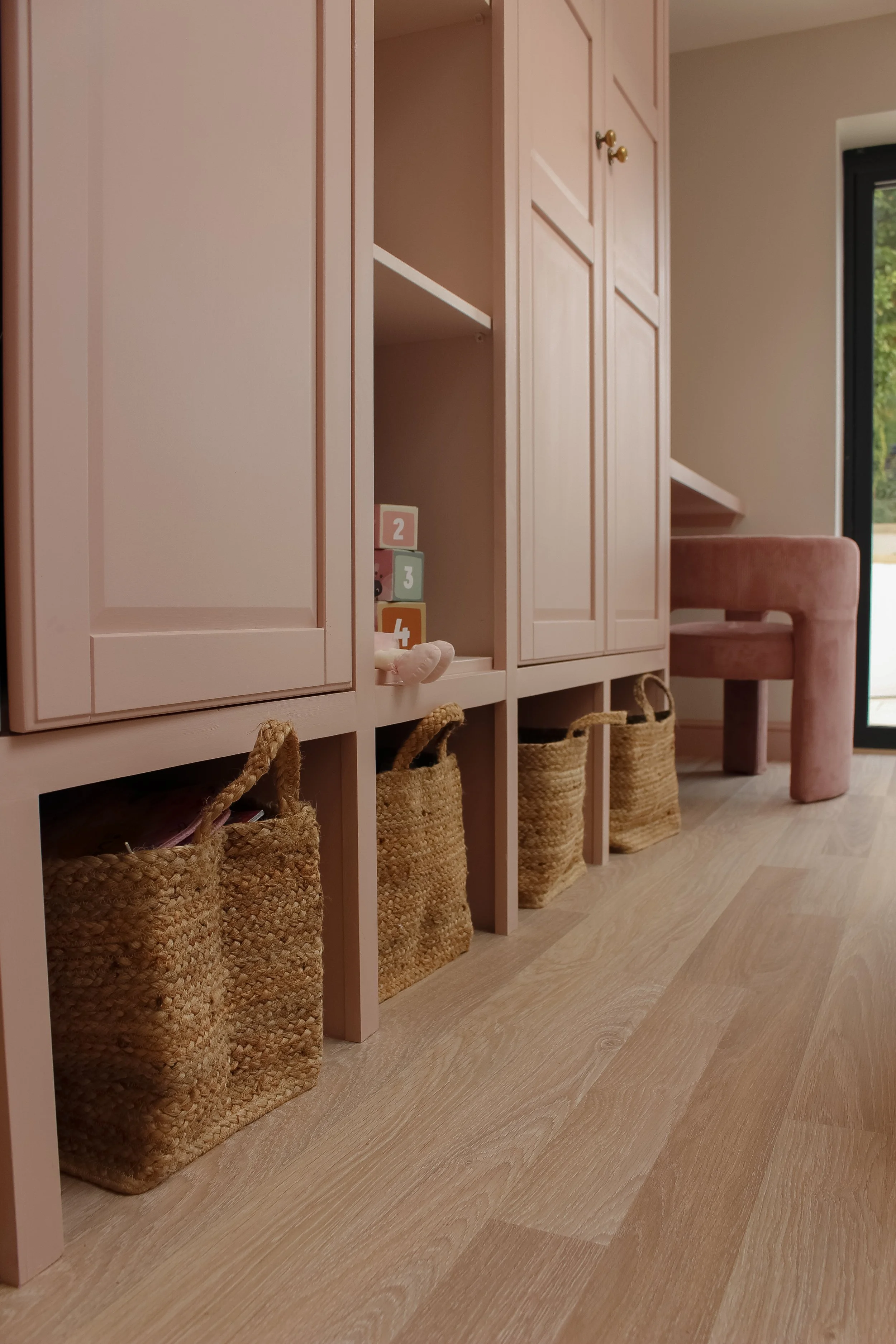 Pink storage cabinet with woven baskets underneath, a velvet pink chair, and a window with a view outside. Playroom designed by Yazzmin Lovelle Interiors.