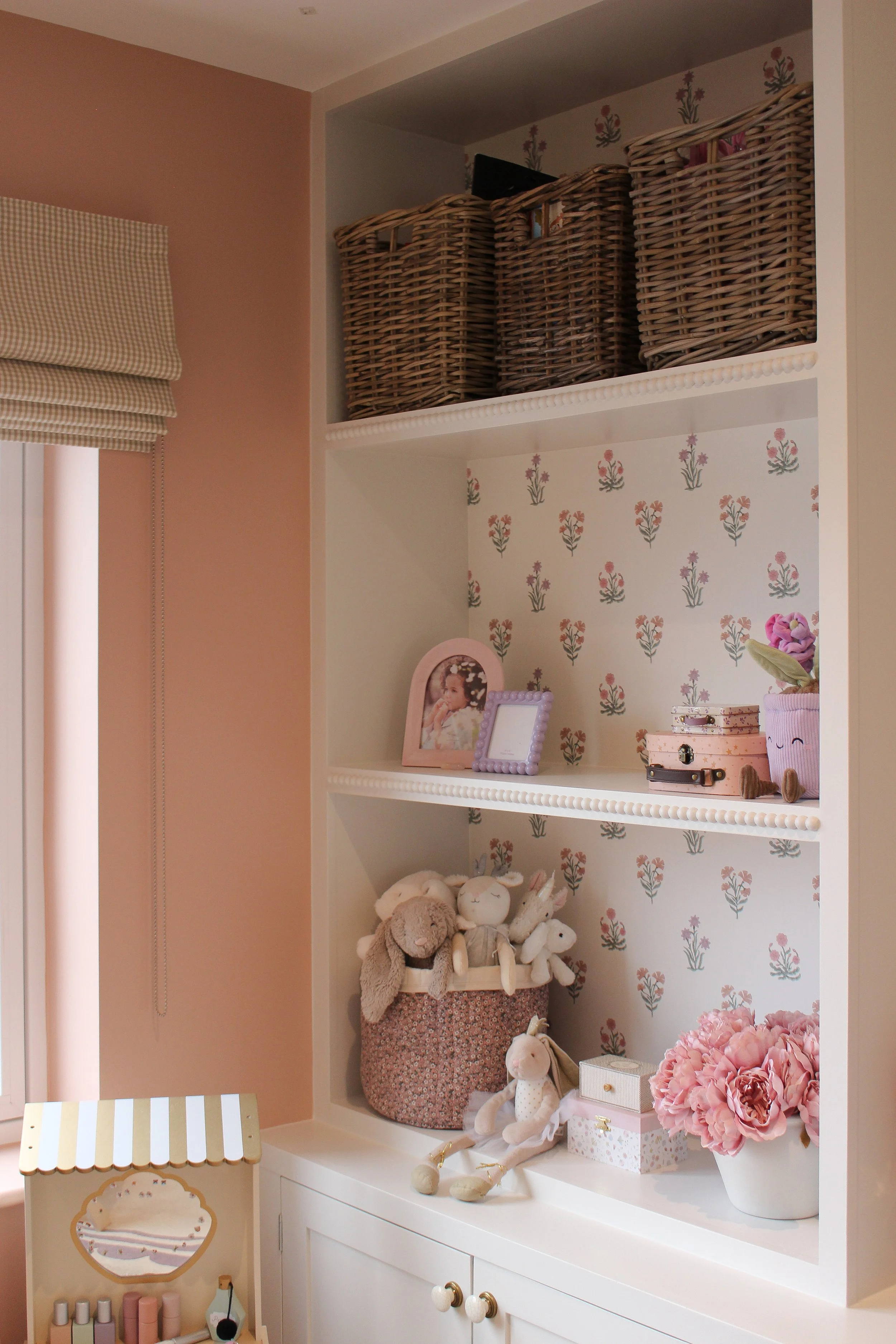 Styled playroom shelf with wicker baskets, photo frames, plush toys, pink flowers, and decorative boxes, against floral wallpaper. Playroom designed by Yazzmin Lovelle Interiors 