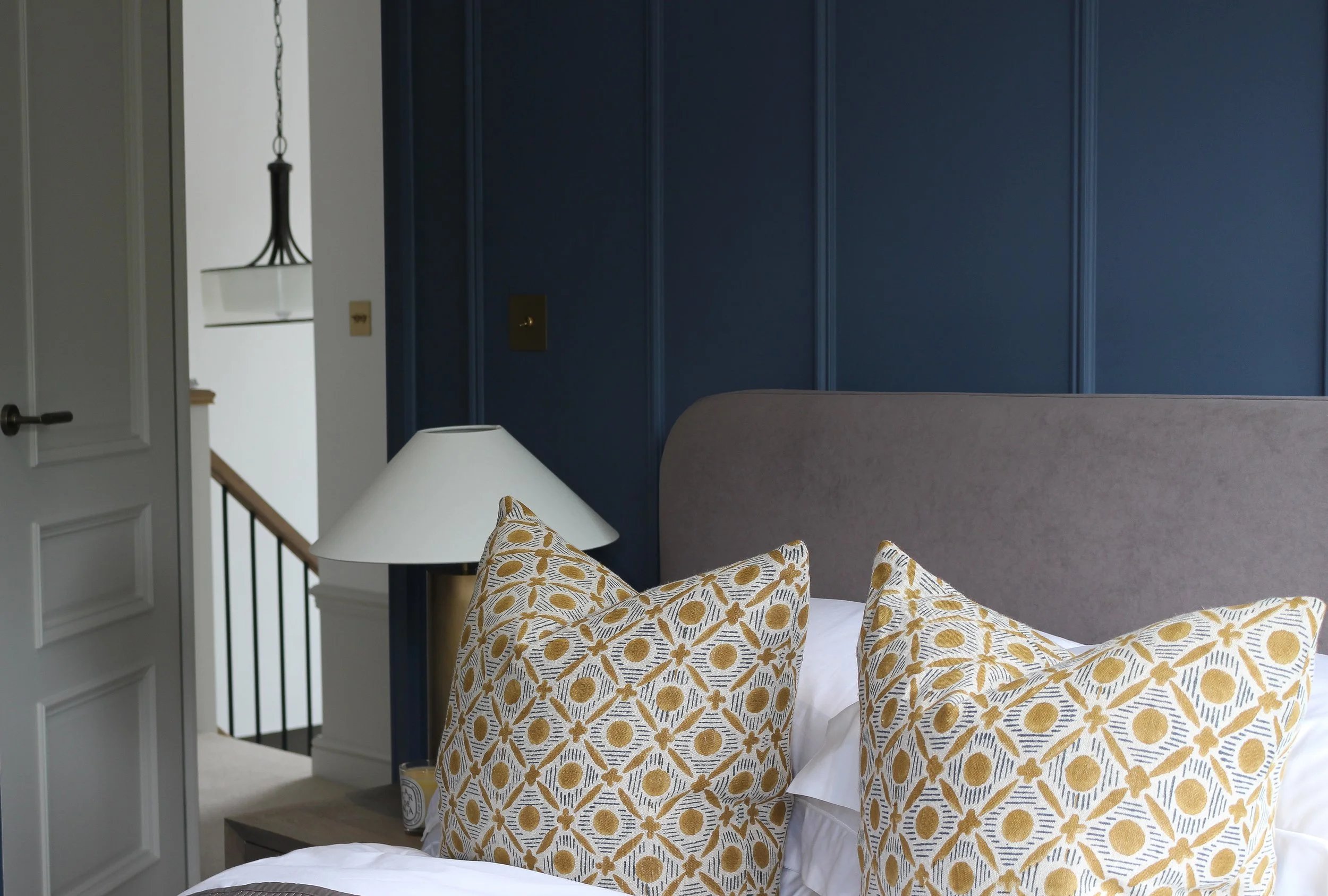 A bedroom with a grey headboard, patterned yellow and white pillows, a wooden bedside table and a white table lamp. The background has a navy blue wall and a partially open white door leading to a staircase area with a hanging light fixture.