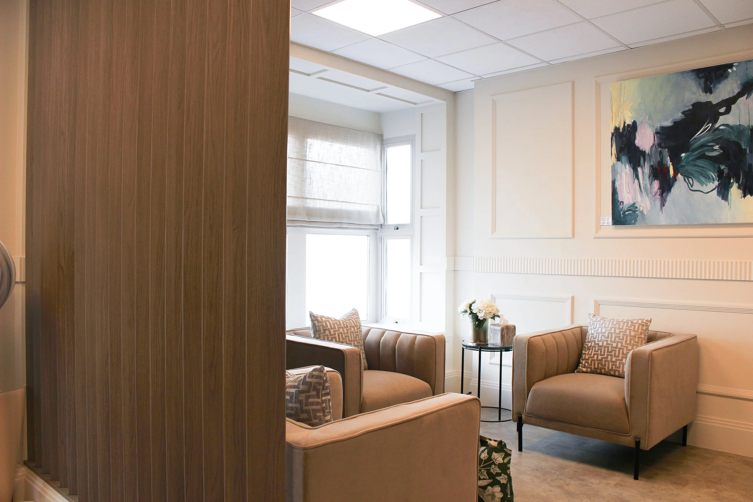 A clinic sitting area with beige armchairs, patterned throw pillows, a small round black side table with a flower vase, and large windows with beige blinds. A colourful abstract painting hangs on the white wall.