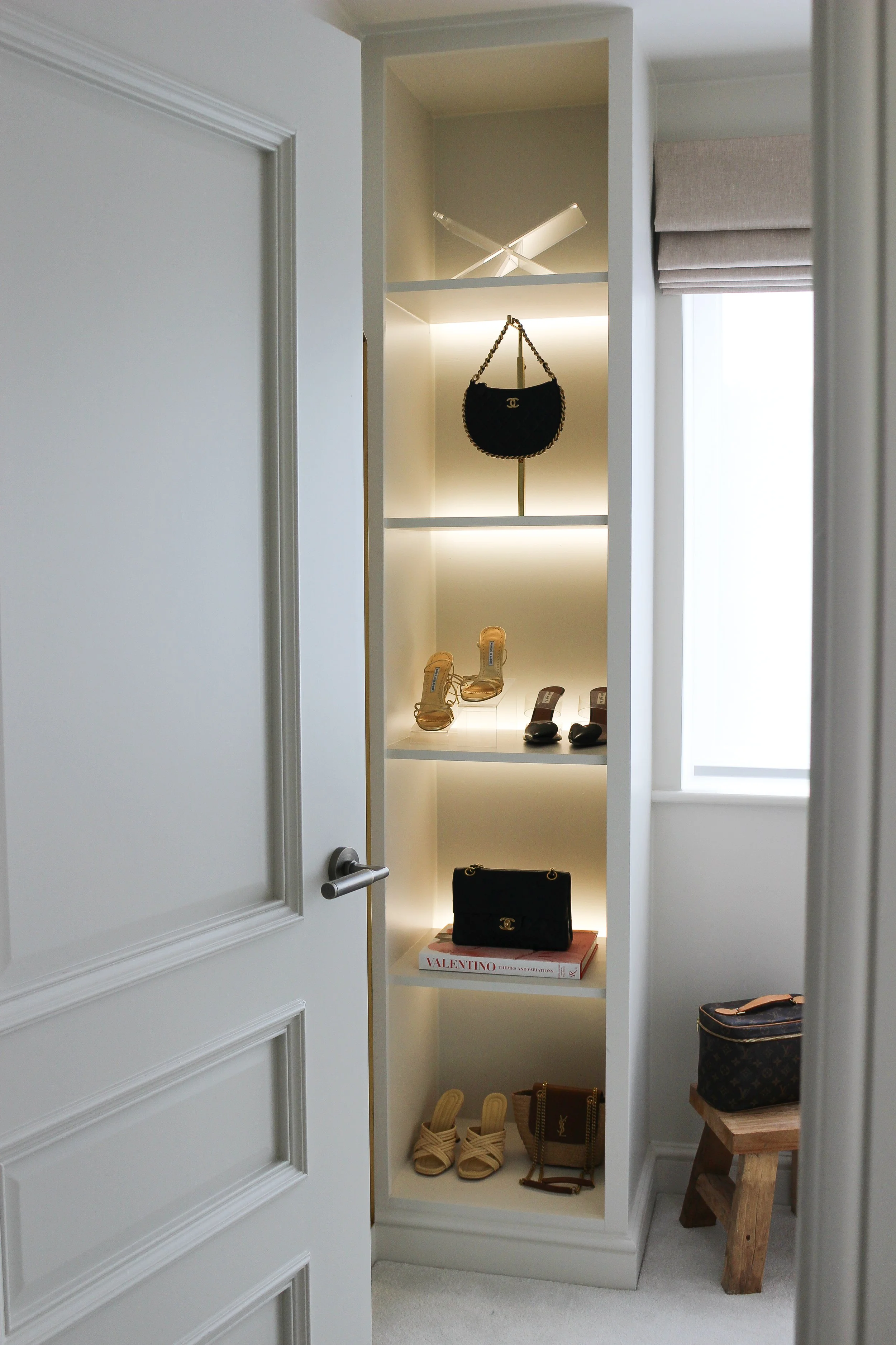 View of a built in white display shelf with illuminated sections showcasing designer handbags, shoes, and accessories in a bright room next to a window. Dressing room designed by Yazzmin Lovelle Interiors