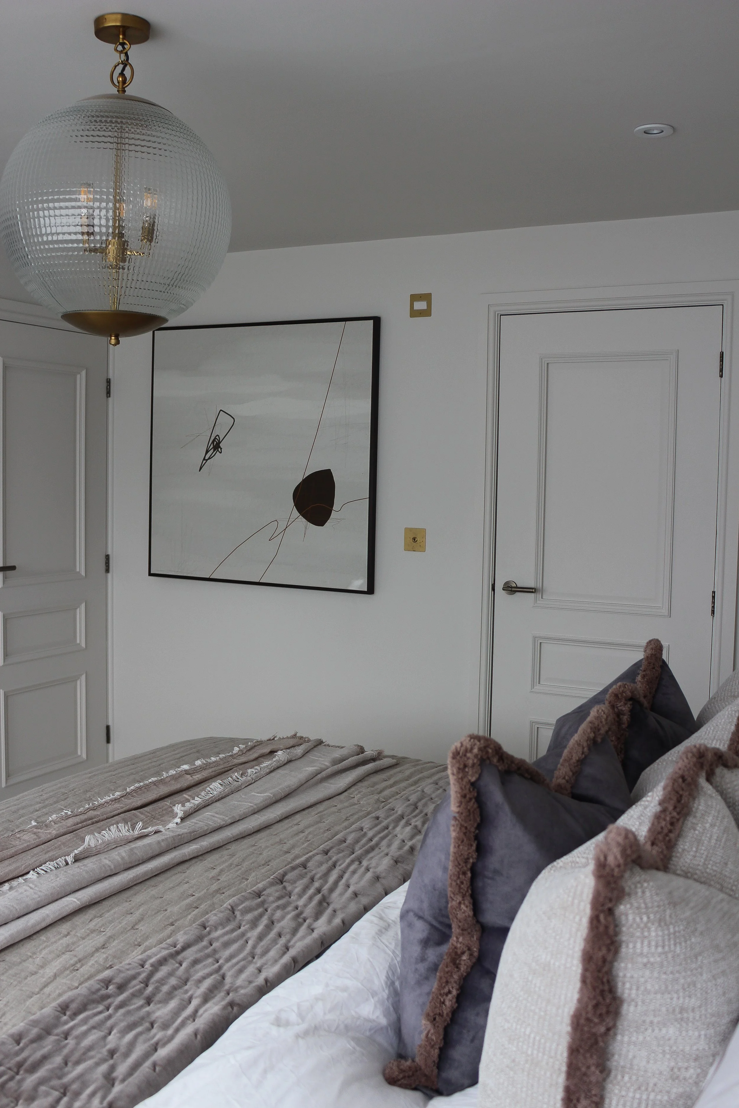 A master bedroom featuring a bed with multiple pillows, a beige bedspread, and artwork on the white wall, with a glass pendant light hanging from the ceiling. Master bedroom designed by Yazzmin Lovelle Interiors