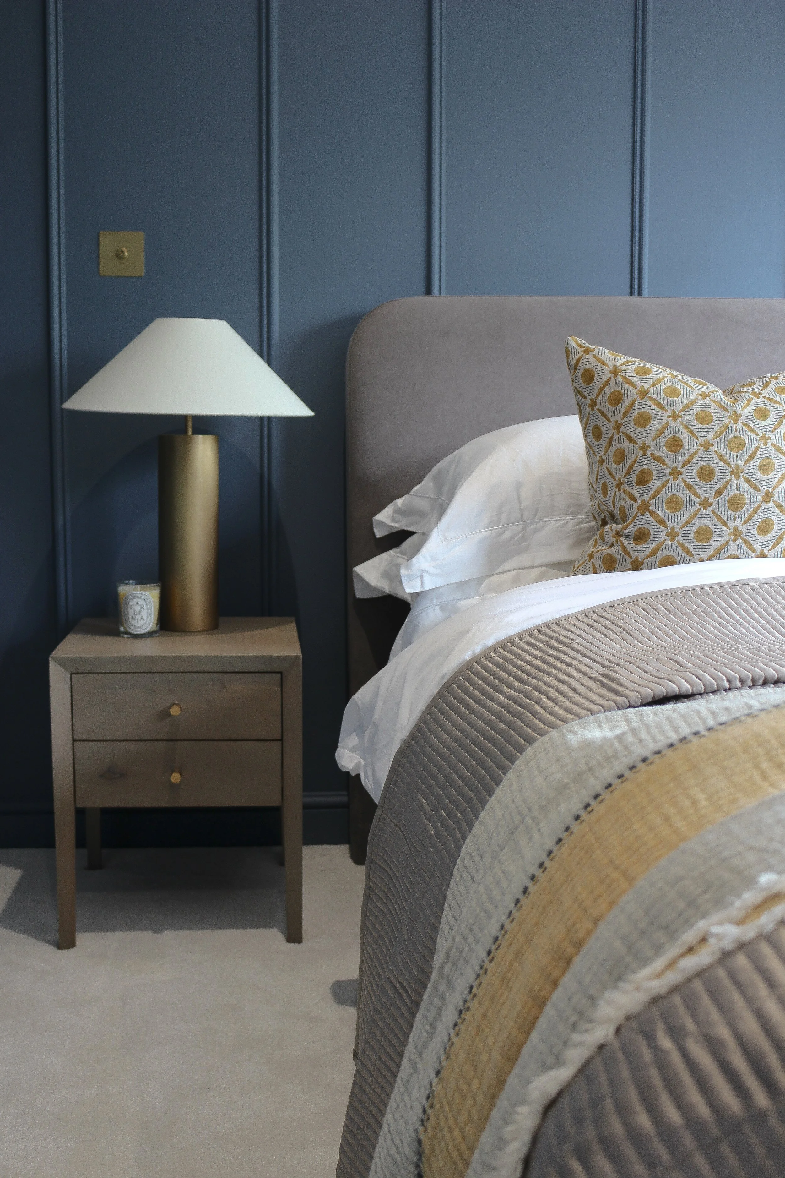 A neatly made bed with white pillows and a patterned decorative pillow, a beige and grey quilt, beside a wooden bedside table with a gold lamp and a small candle, against a blue panelled wall.