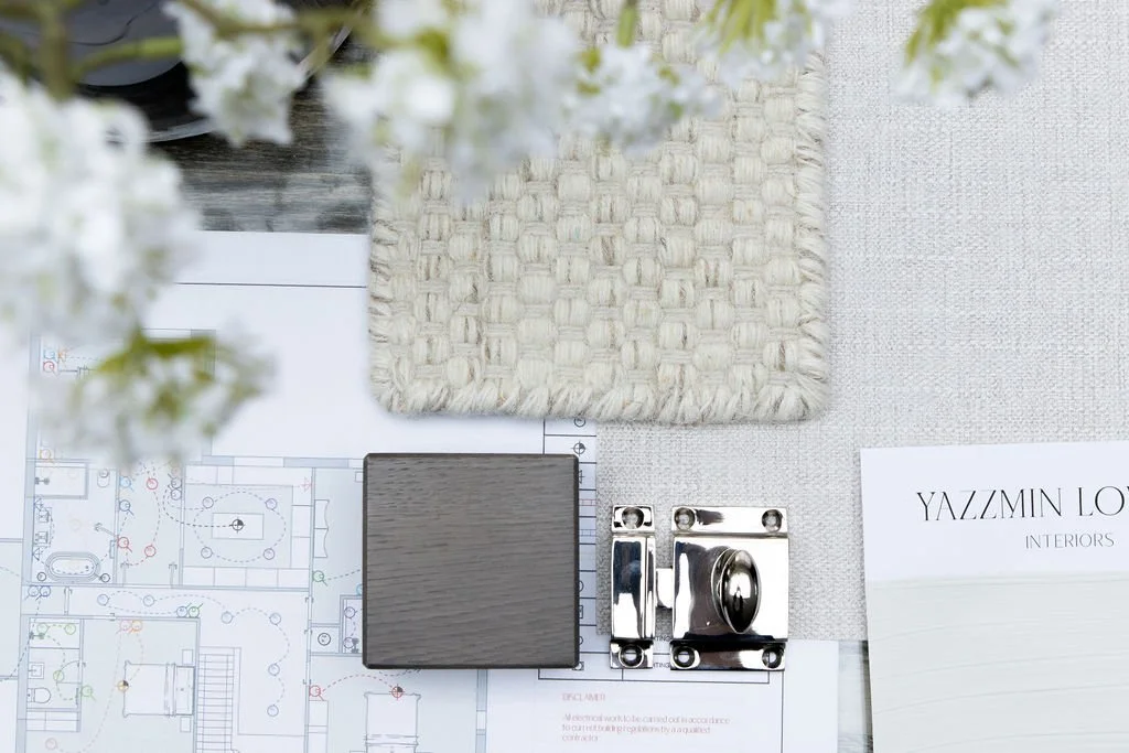 Yazzmin Lovelle Interiors interior design samples, including a woven fabric swatch, a small dark wood sample, a silver hardware piece, a floral arrangement, and an interior design electrical plan on a white surface.