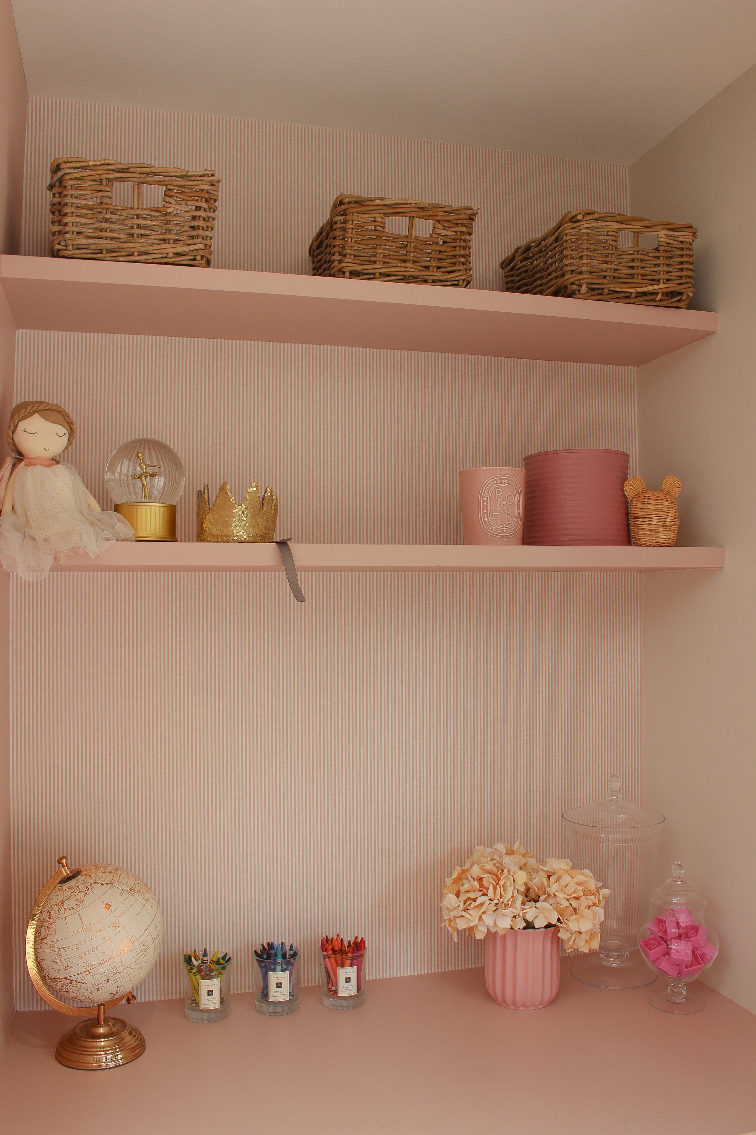 stripe pink wallpaper holding wicker baskets, a doll, a snow globe, a gold crown, pink containers, and a small teddy bear figure; on the table below are coloured pencils, a flower arrangement, glass jars. 