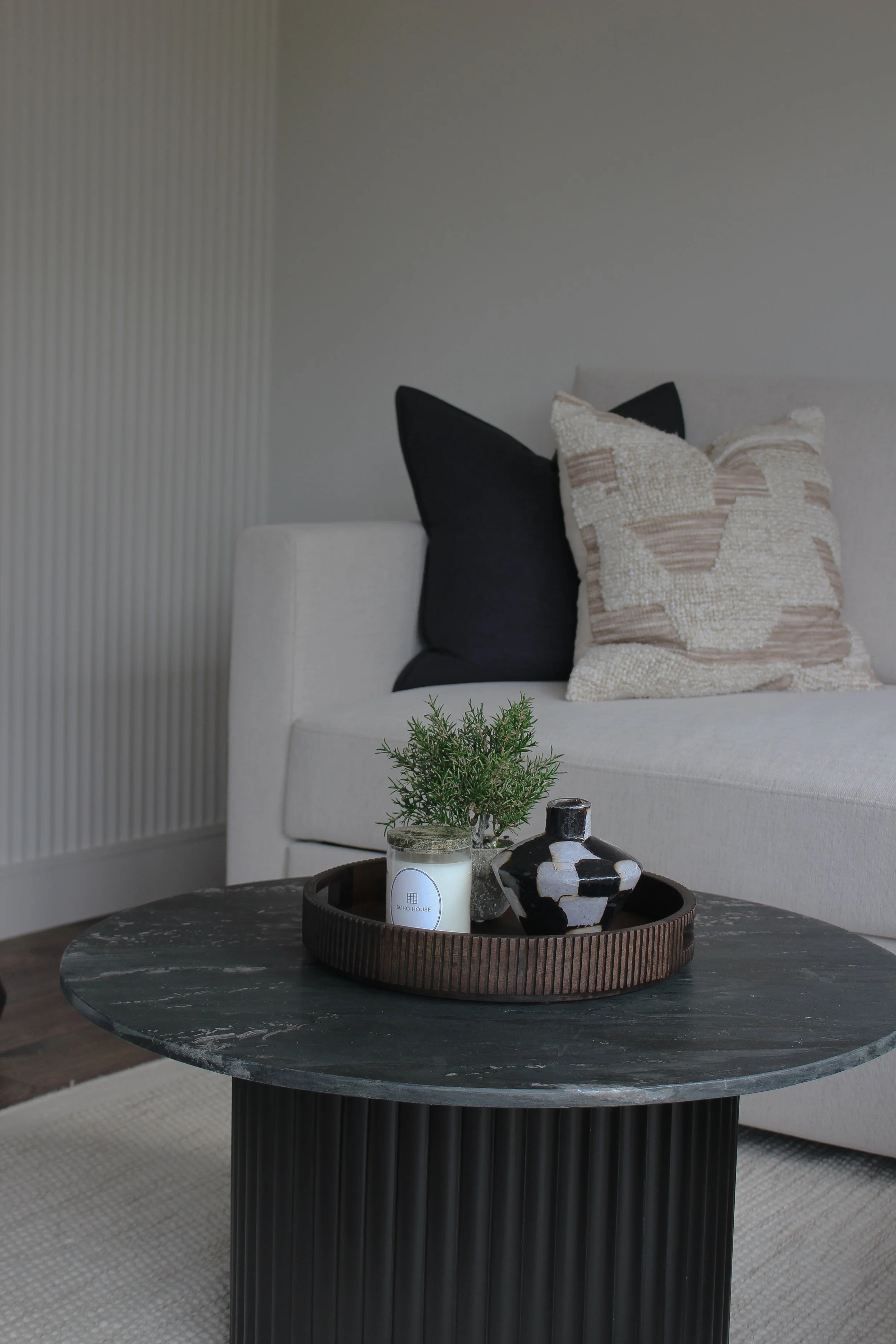 Office with a white sofa, beige and black pillows, a round black coffee table with a tray, plant, candle, and decorative vase on it. Garden office designed by Yazzmin Lovelle Interiors.