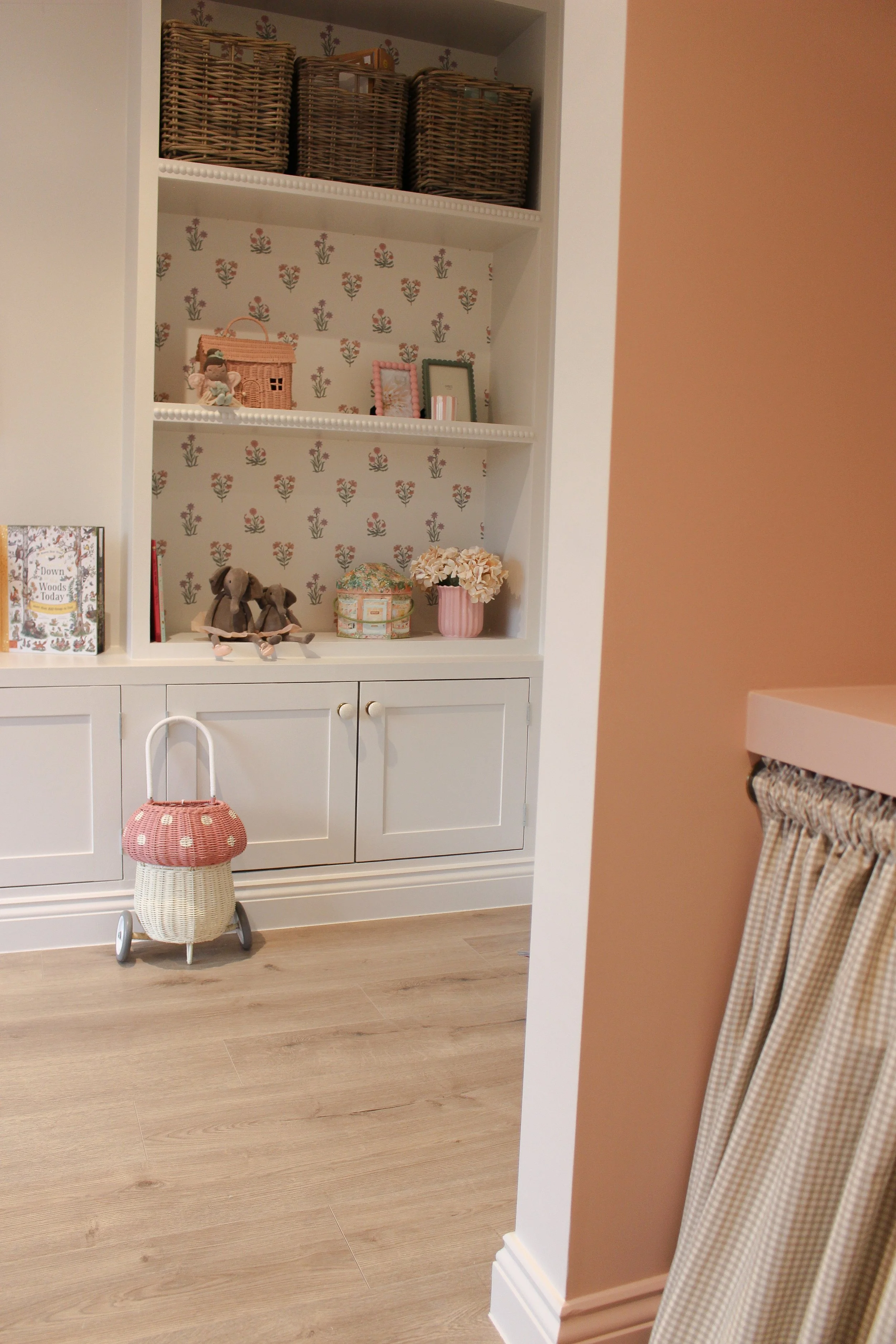A white built in bookshelf in a playroom with floral wallpaper, decorated with small toys, books, and floral arrangements, a wicker trolly at the base, and a peach coloured wall on the right with a gingham curtain.