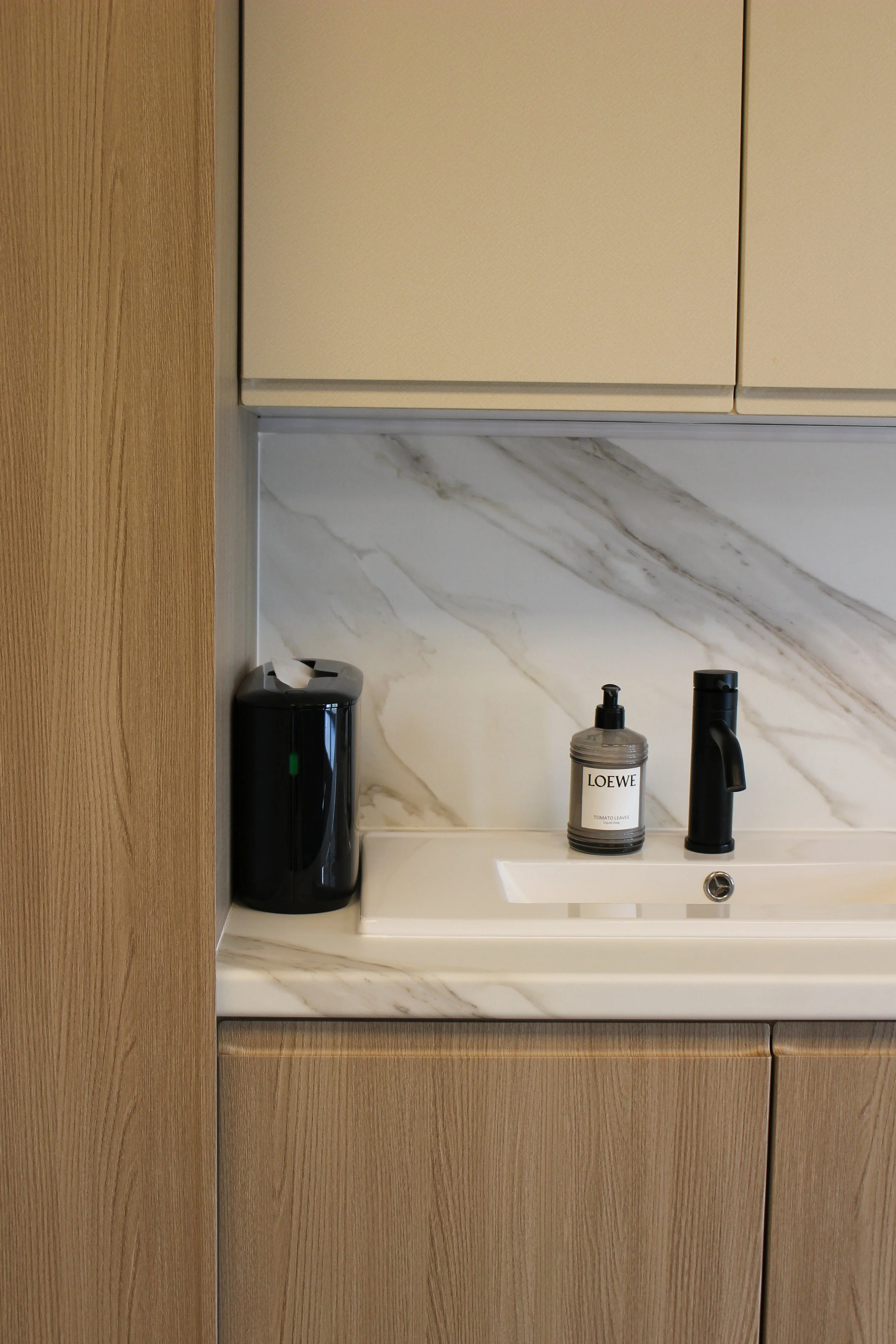 Clinic countertop with a black tissue dispenser, a soap pump labeled 'LOEWE', and a black tap, against a marble backsplash and wooden cabinets. Clinic designed by Yazzmin Lovelle Interiors