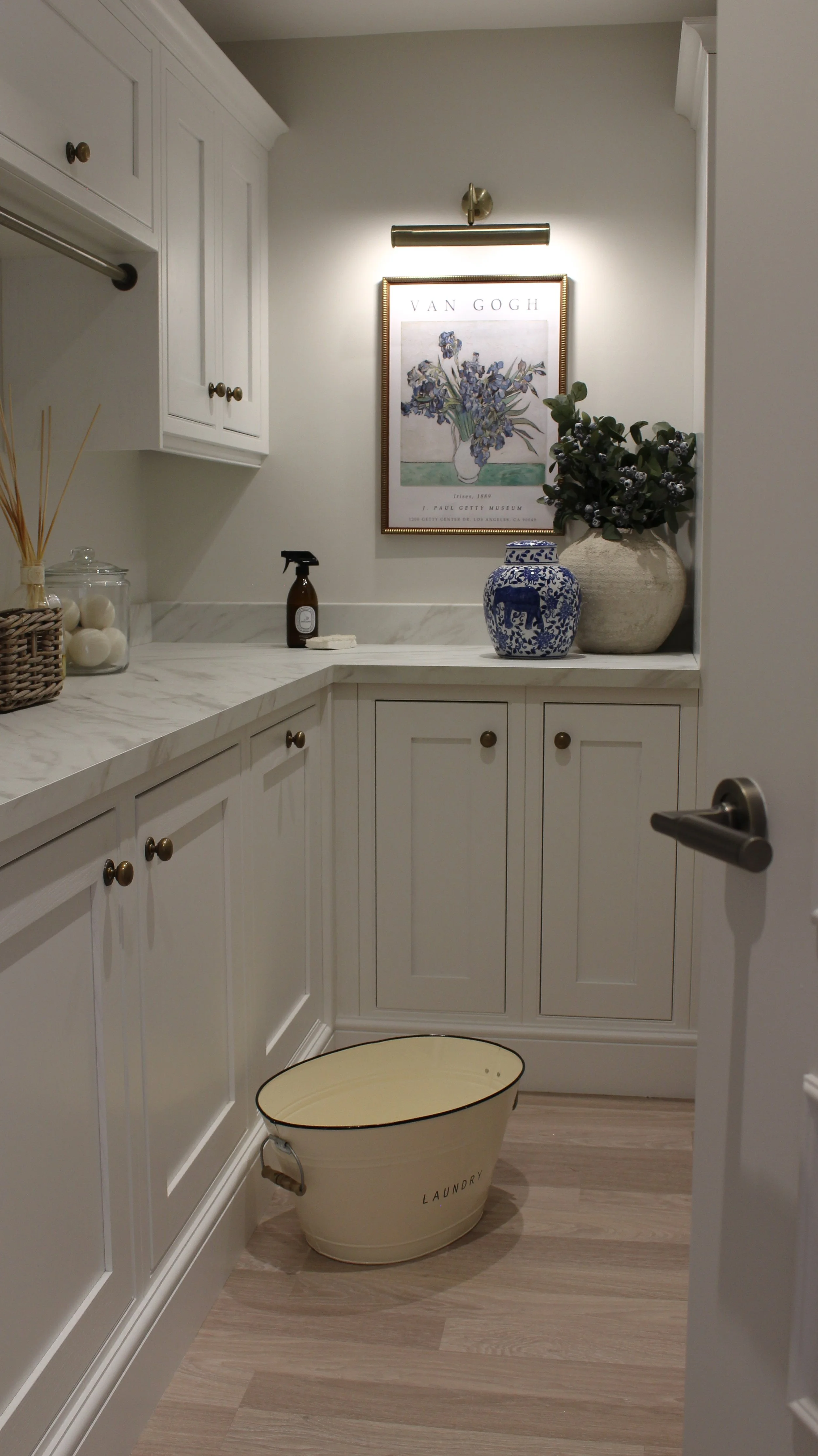 A laundry room with white cabinets, marble countertops, a framed Van Gogh art print, a blue and white ceramic jar, a large beige vase with greenery, a small spray bottle, a glass jar with white balls, a reed diffuser, and a beige laundry basket on a 