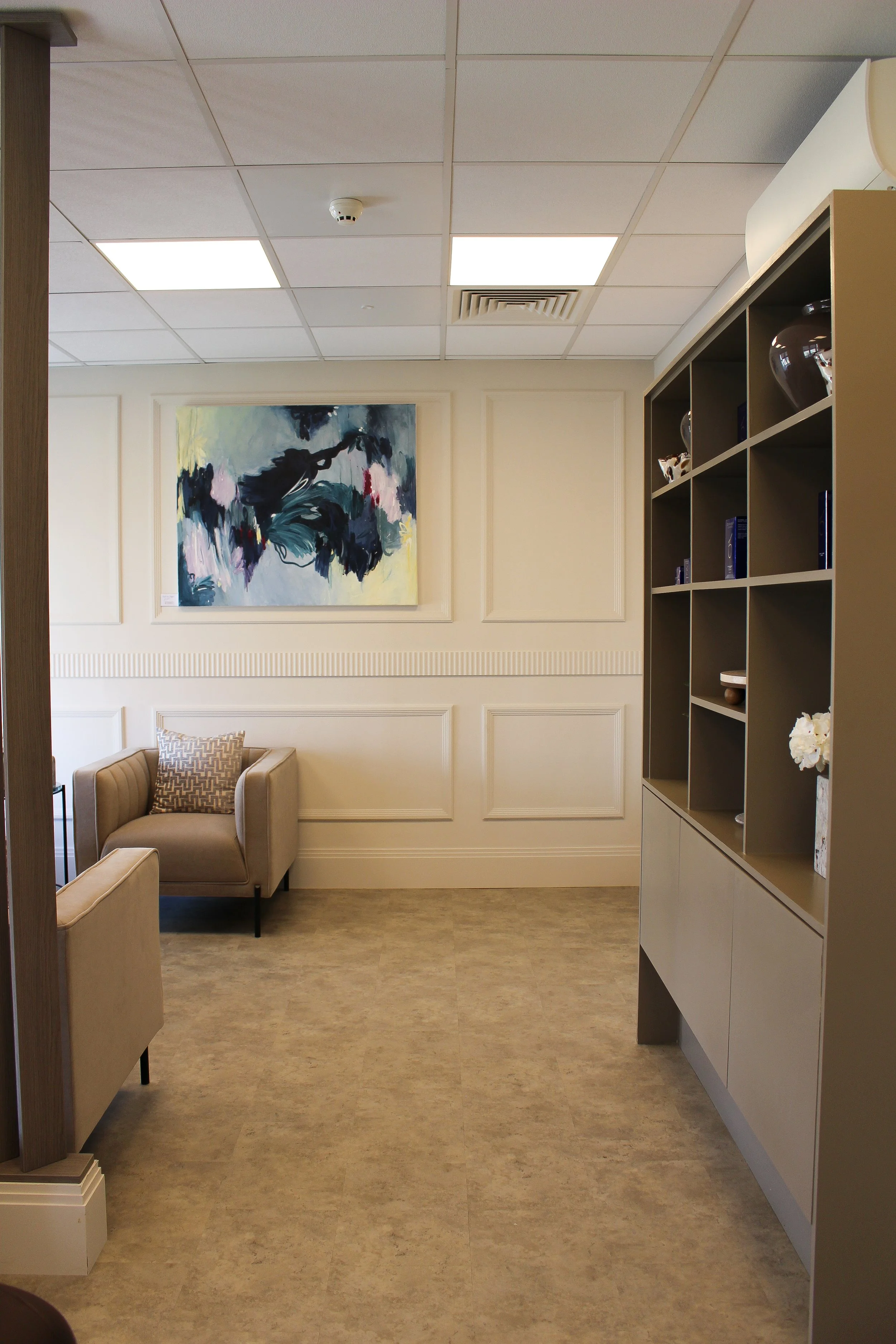 A luxury seating room with beige armchairs, a patterned pillow, abstract artwork on the wall, a beige shelving unit with decorative items, subdued lighting, and a stone tiled floor. Clinic designed by Yazzmin Lovelle Interiors