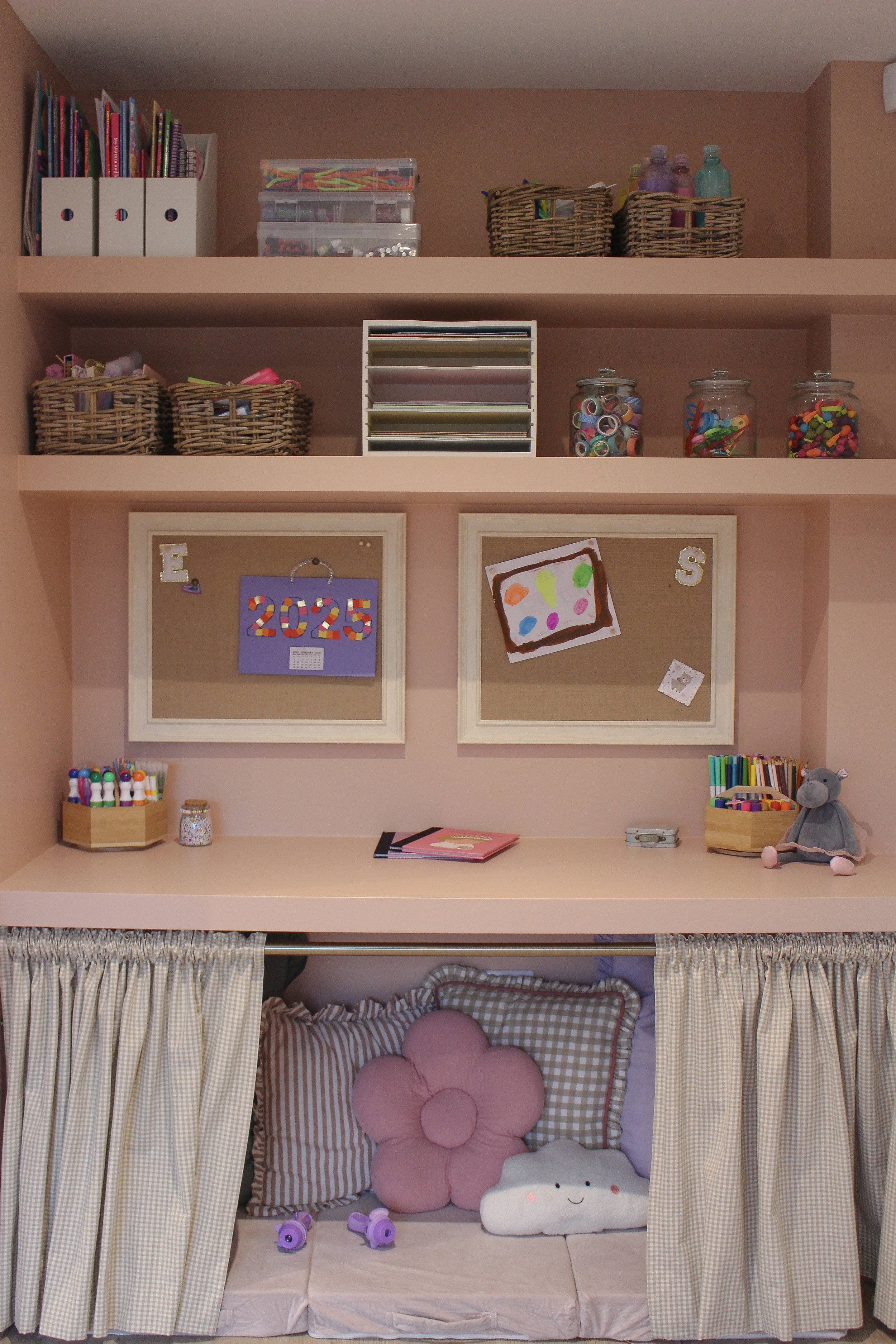 A children's craft and play area with shelves containing art supplies, decorative jars, and organised boxes. wall has a notice board with children's art. Below is a children's playden with fun cushions. Playroom designed by Yazzmin Lovelle Interiors 