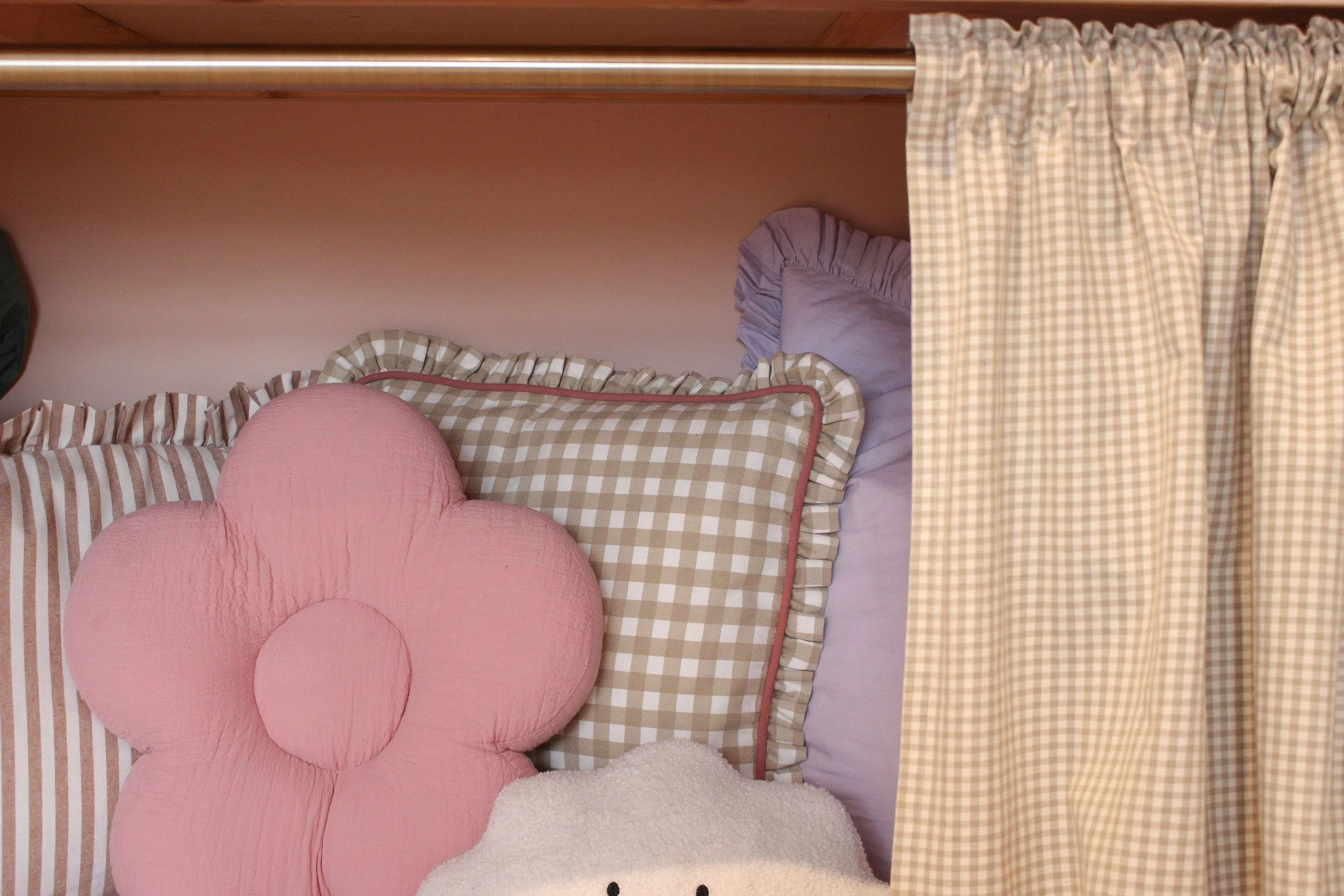 Arrangement of decorative pillows in pastel colours including a pink flower shaped pillow, checkered pillows with ruffled edges, and a cream textured pillow, behind a beige and white gingham curtain over a playroom den