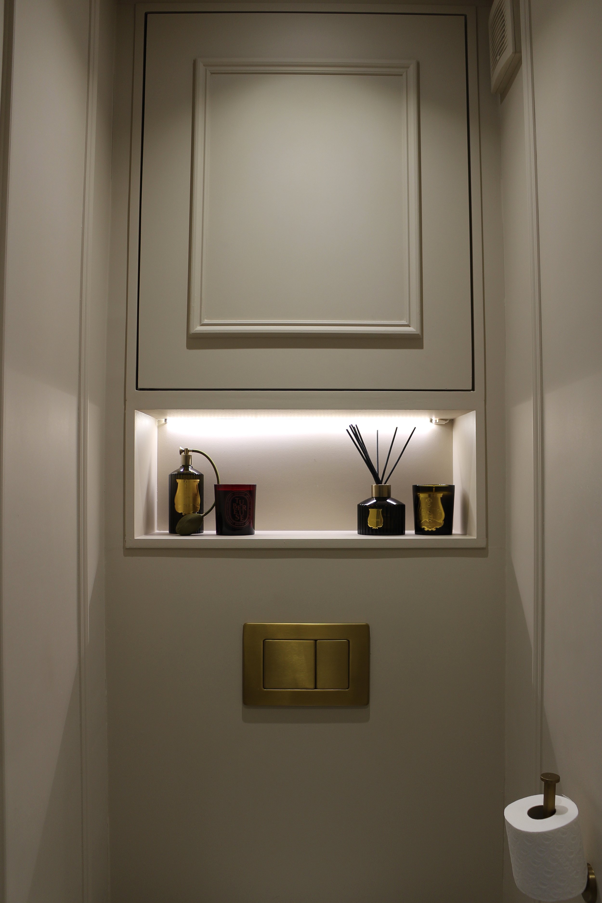A small built-in wall niche with decorative bottles, a reed diffuser, and candles, illuminated by soft lighting, surrounded by white walls and a gold flush button on the wall below. Downstairs toilet designed by Yazzmin Lovelle Interiors 