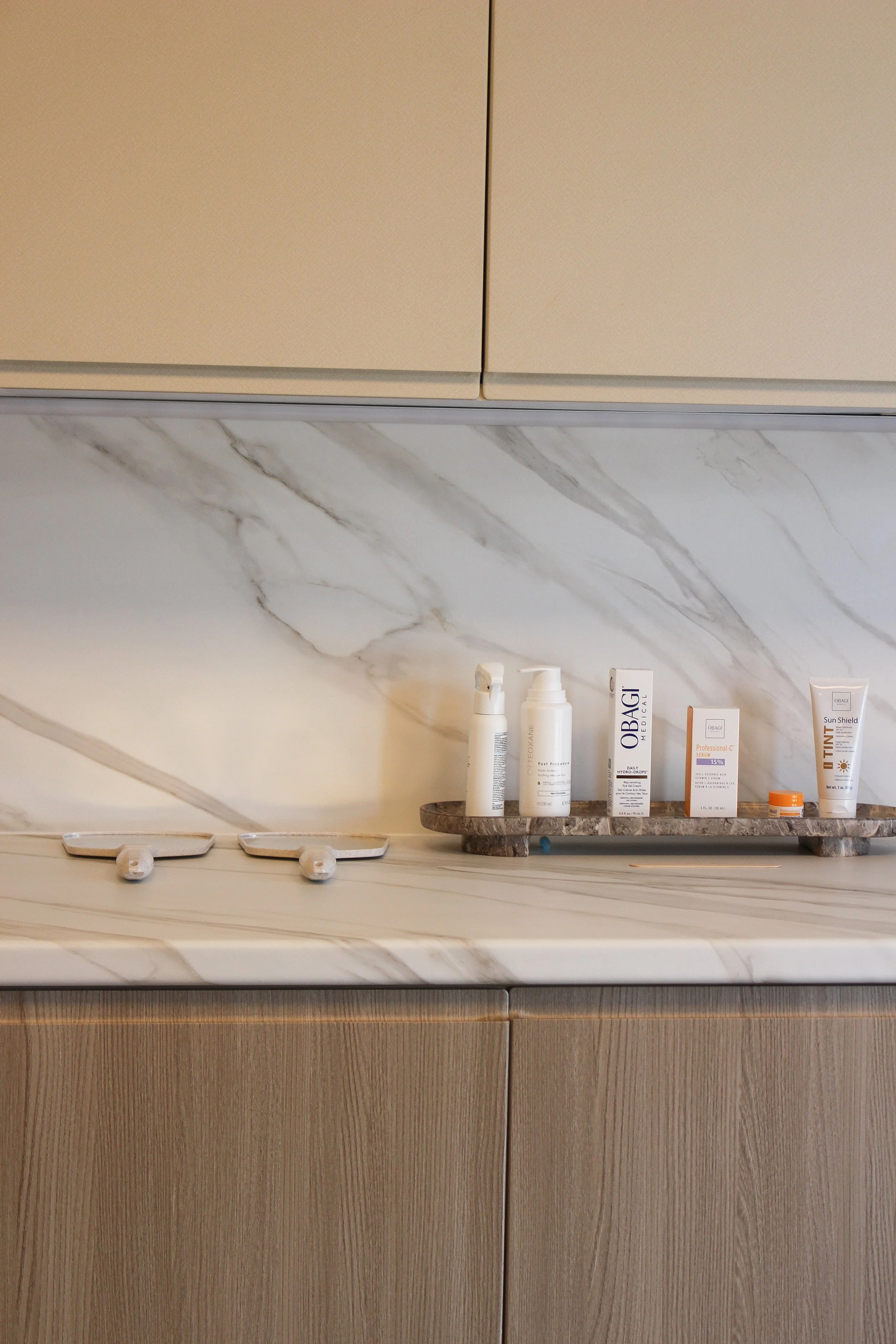 A marble countertop with skincare and sun protection products on a wooden tray and two small wall hooks in a luxury clinic. Clinic designed by Yazzmin Lovelle Interiors