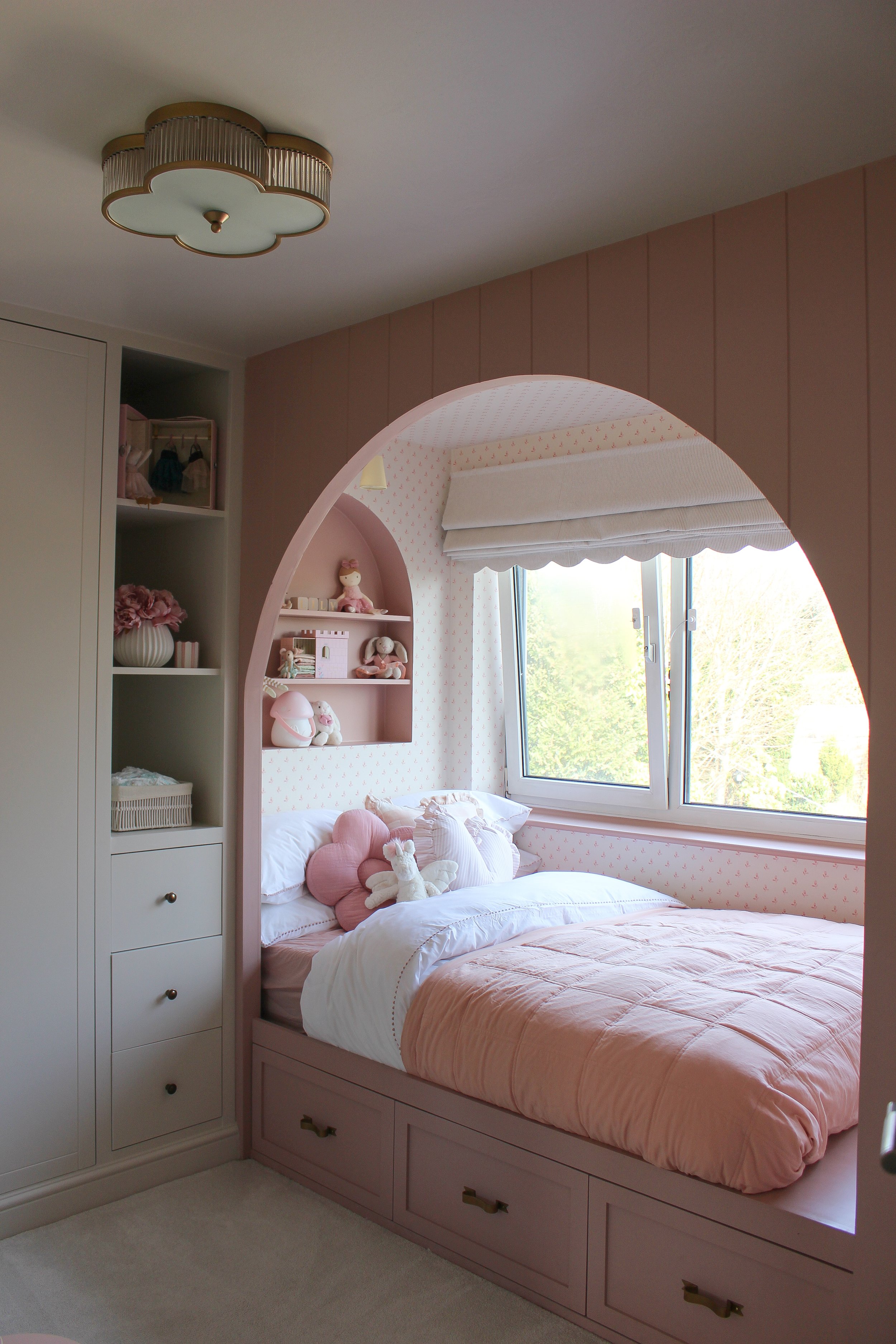A girls bedroom with pink and white decor, a built in bed with arched entrance with plush pillows and stuffed animals, a window with a view of trees, and built-in shelves with decorative items. Little girls room designed by Yazzmin Lovelle Interiors.