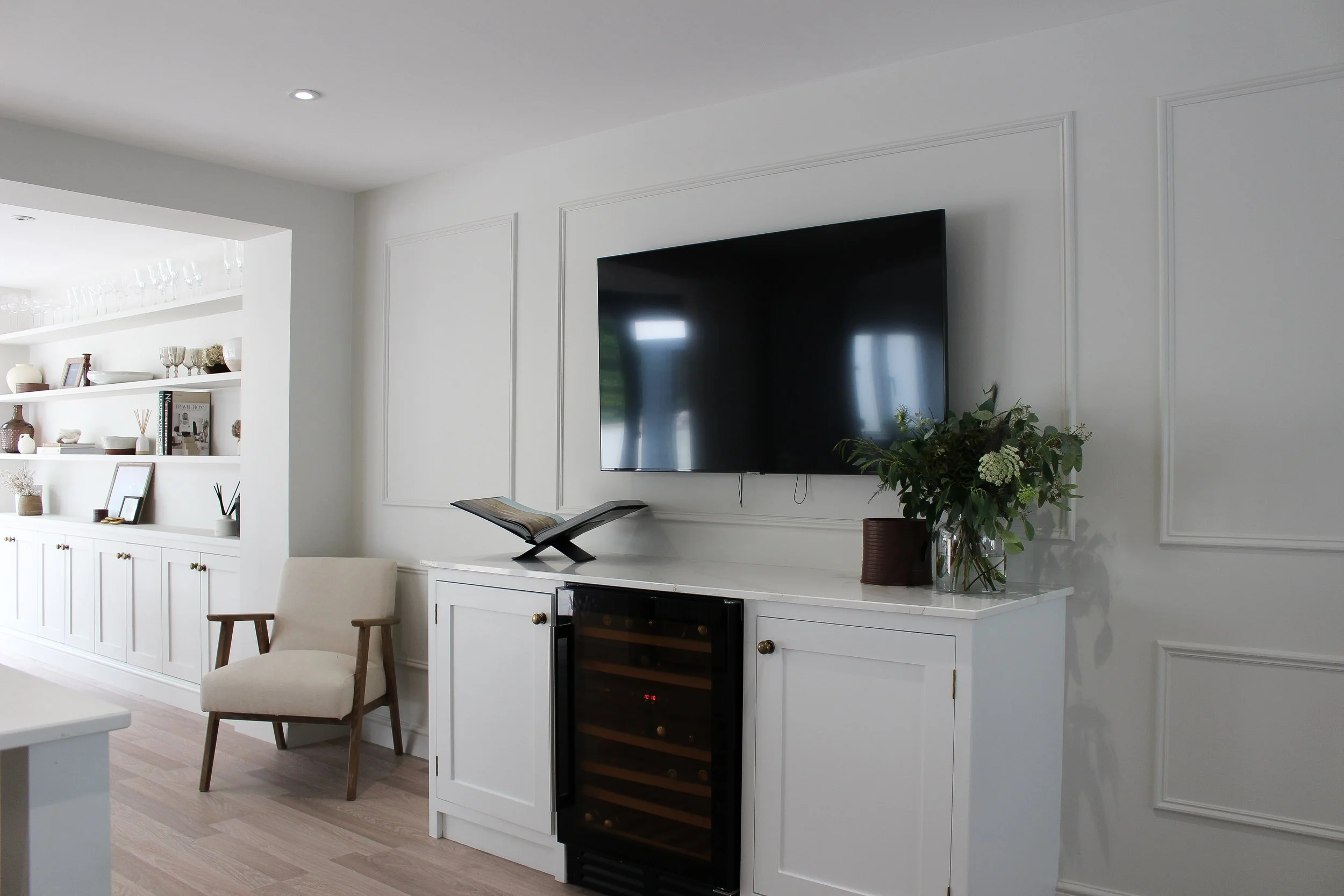 Living room with a wall-mounted flat-screen TV, white cabinetry with a wine cooler, a vase with greenery, an open book, and a beige armchair. Built-in shelves on the adjacent wall hold decorative items and framed pictures.