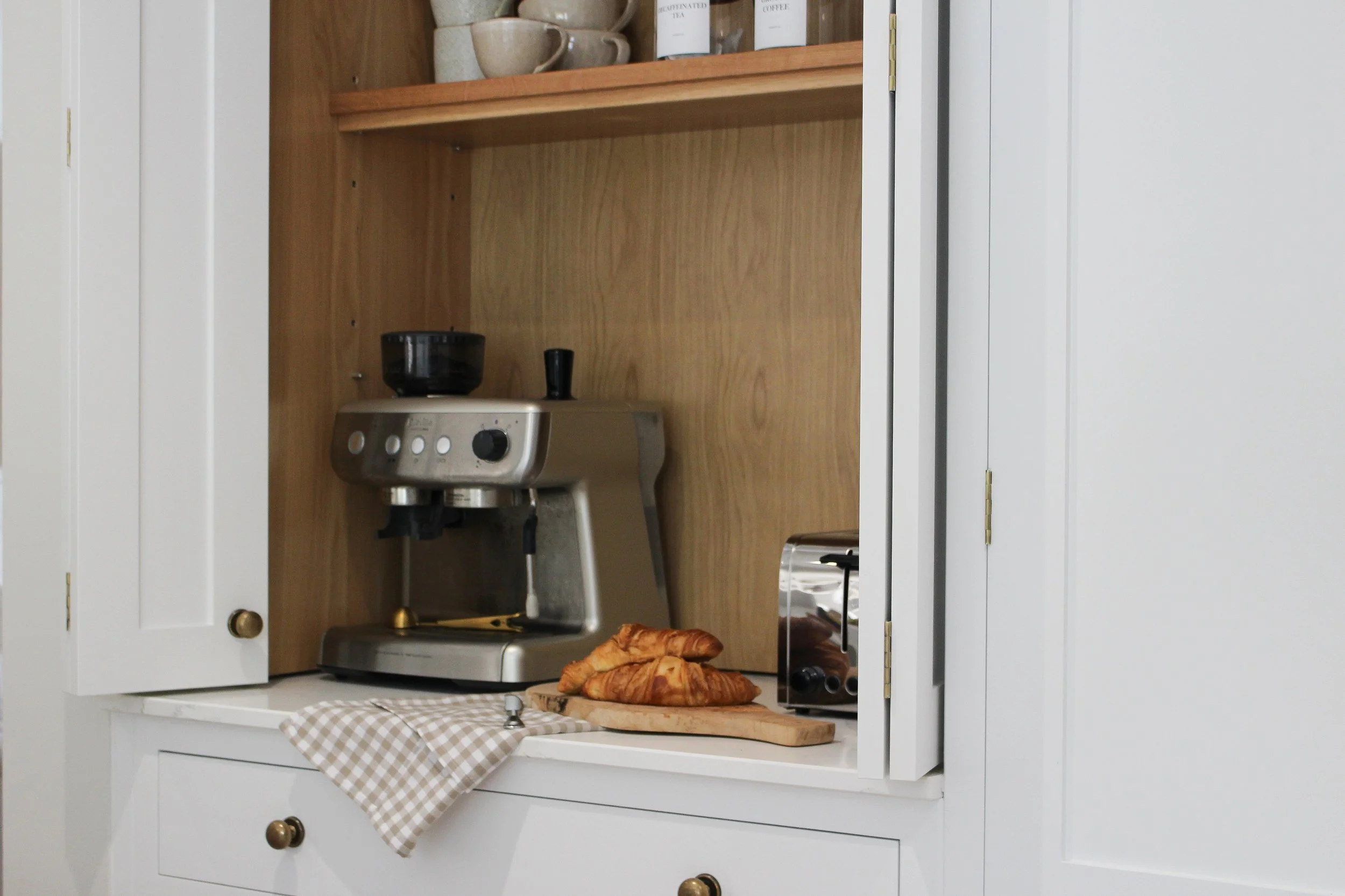 Coffee station with an espresso machine, croissants on a wooden cutting board, a toaster, and ceramic cups on wooden shelves.