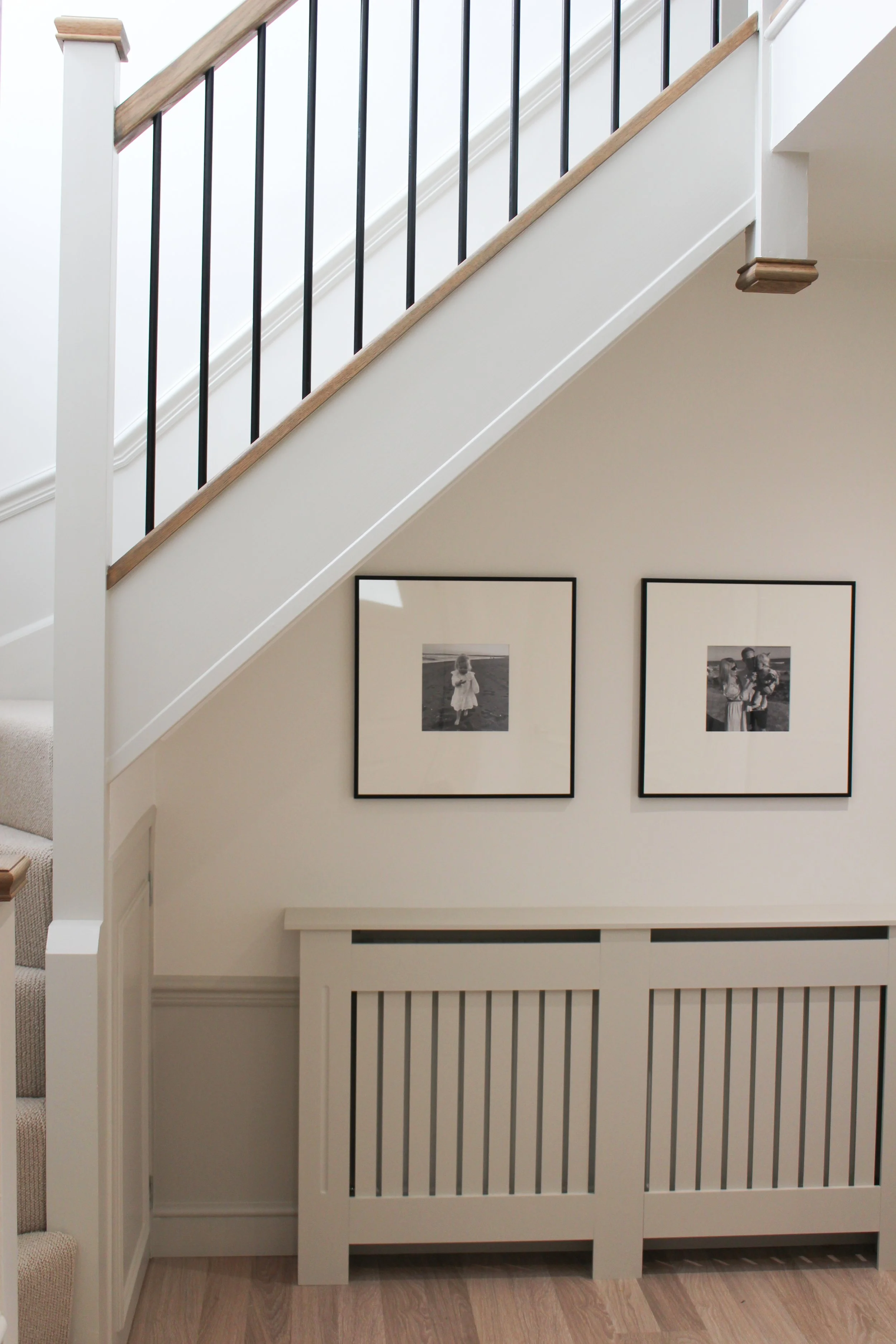 Interior view of a staircase with white walls and black and wooden handrails, decorated with two framed black and white photographs on the wall underneath the staircase.