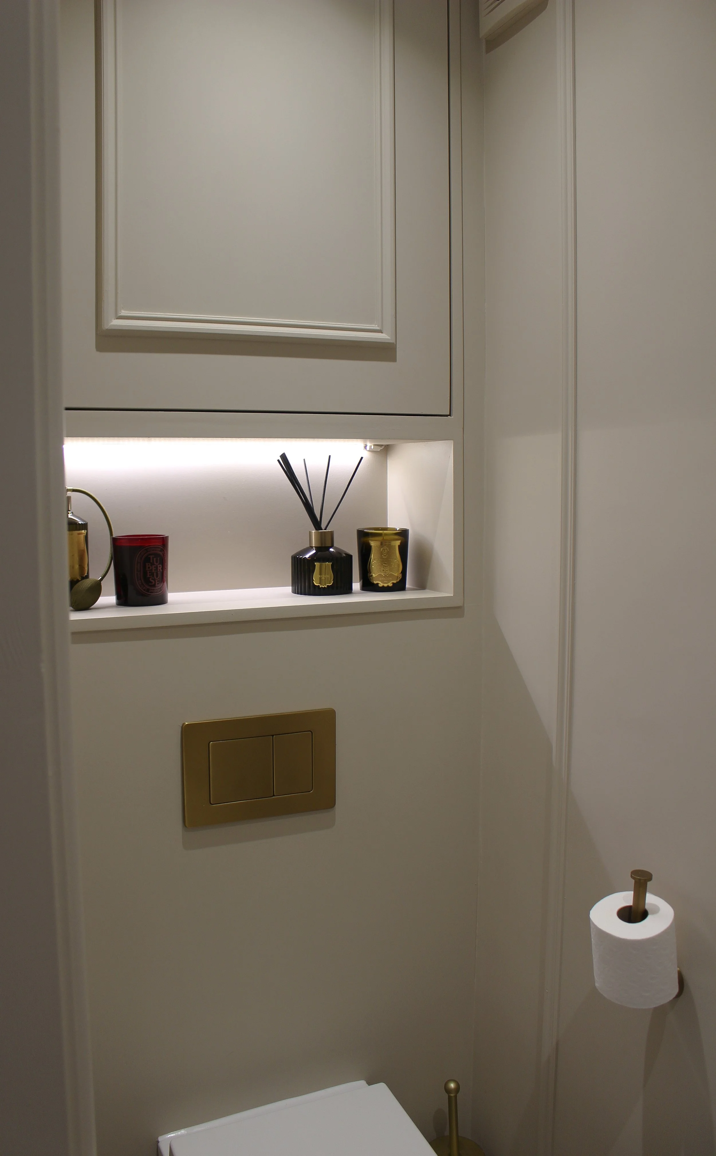A small styled downstairs bathroom with a cream wall, a gold flush plate, a toilet paper roll on a holder, and a shelf with decorative candles and a reed diffuser. Downstairs toilet designed by Yazzmin Lovelle Interiors 