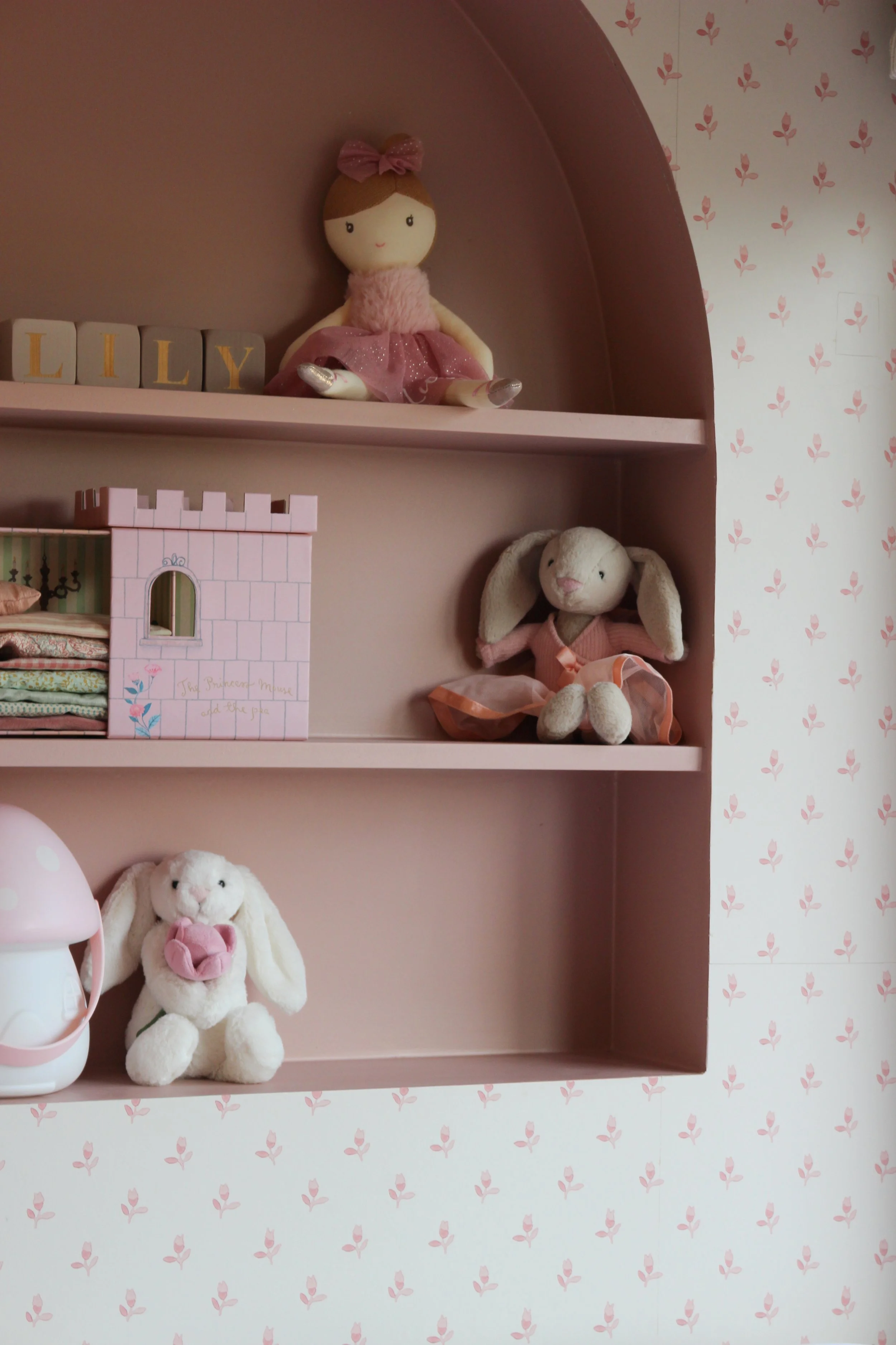 Traditional wallpapered wall with, pink arched shelf with plush toys and decorative items, including dolls and bunny plushies, against pink and floral wallpaper. Girls bedroom designed by Yazzmin Lovelle Interiors