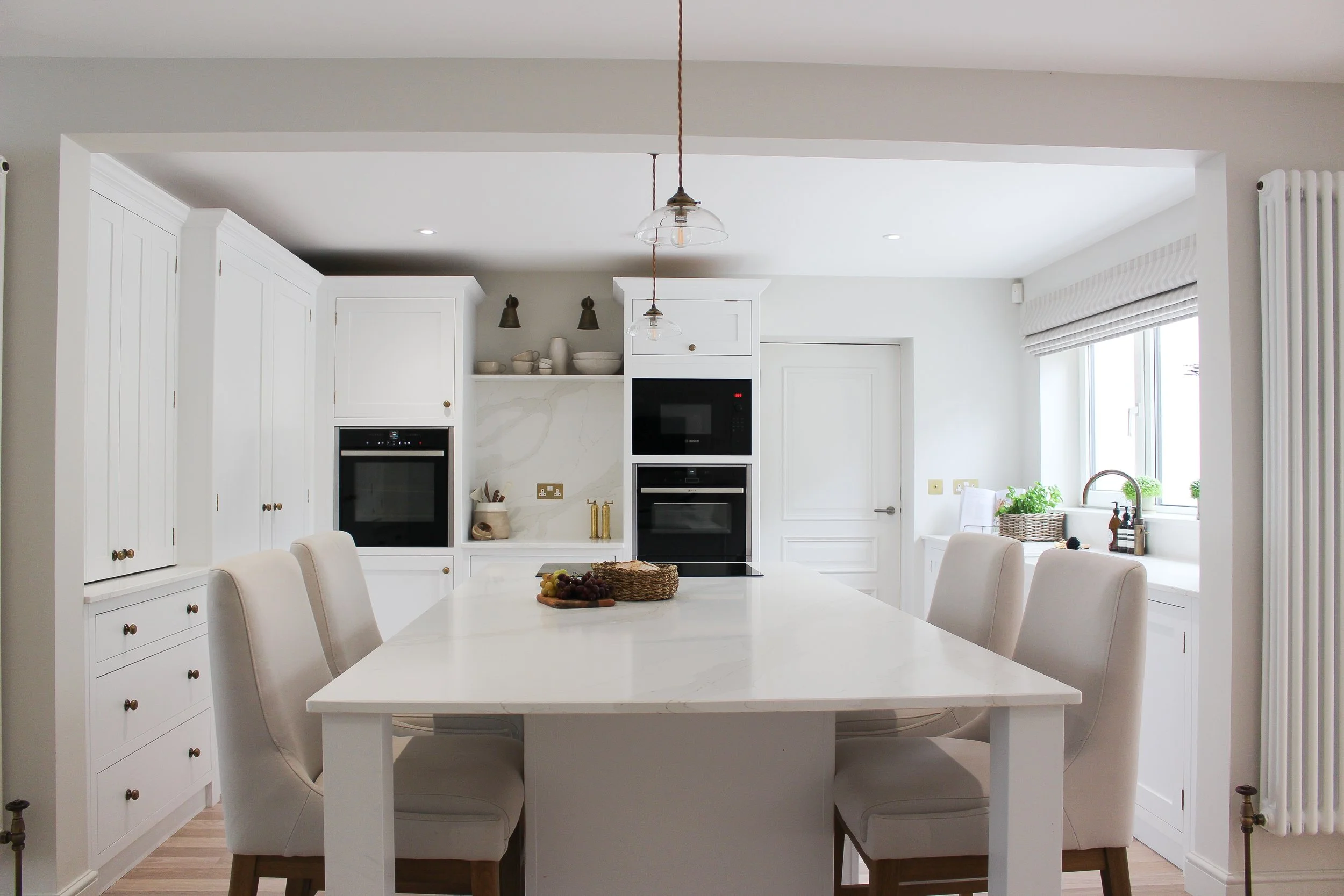 Bright white kitchen with large island, surrounded by white cabinetry, built-in oven and microwave, window with blinds, and beige chairs around the island.