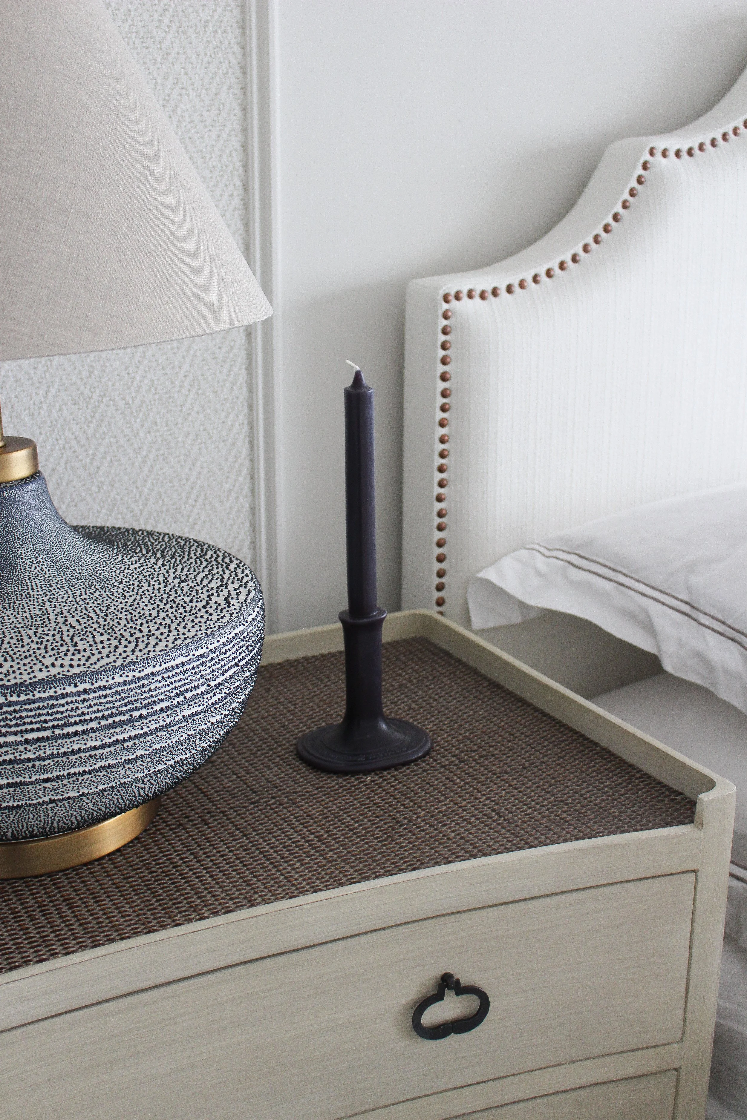 A bedside table with a textured table lamp, a black candle in a holder, and a white headboard with studded accents. Master bedroom designed by Yazzmin Lovelle Interiors