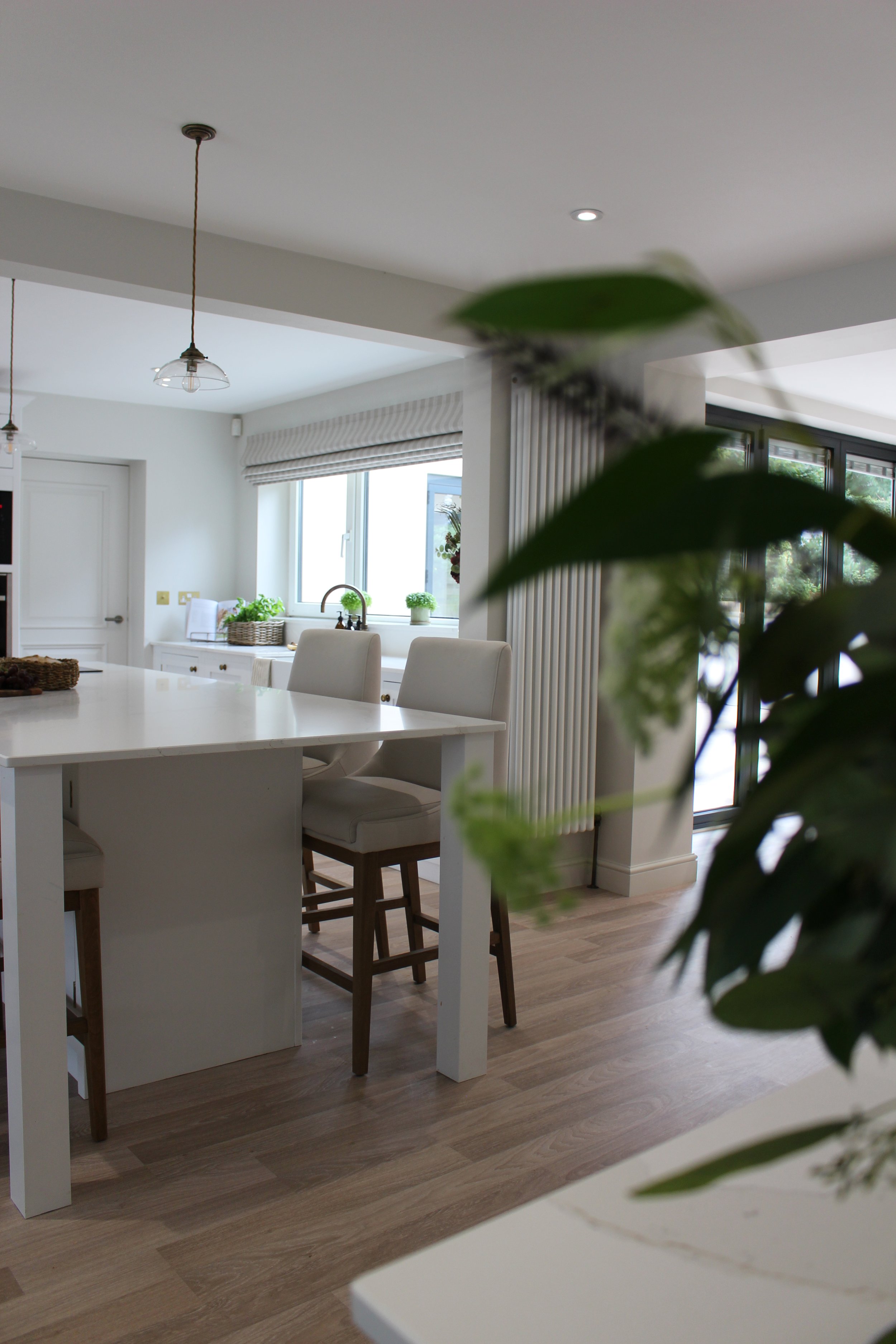 A bright, traditional kitchen and dining area with a white island, beige chairs, wooden flooring, and large windows with outdoor views.