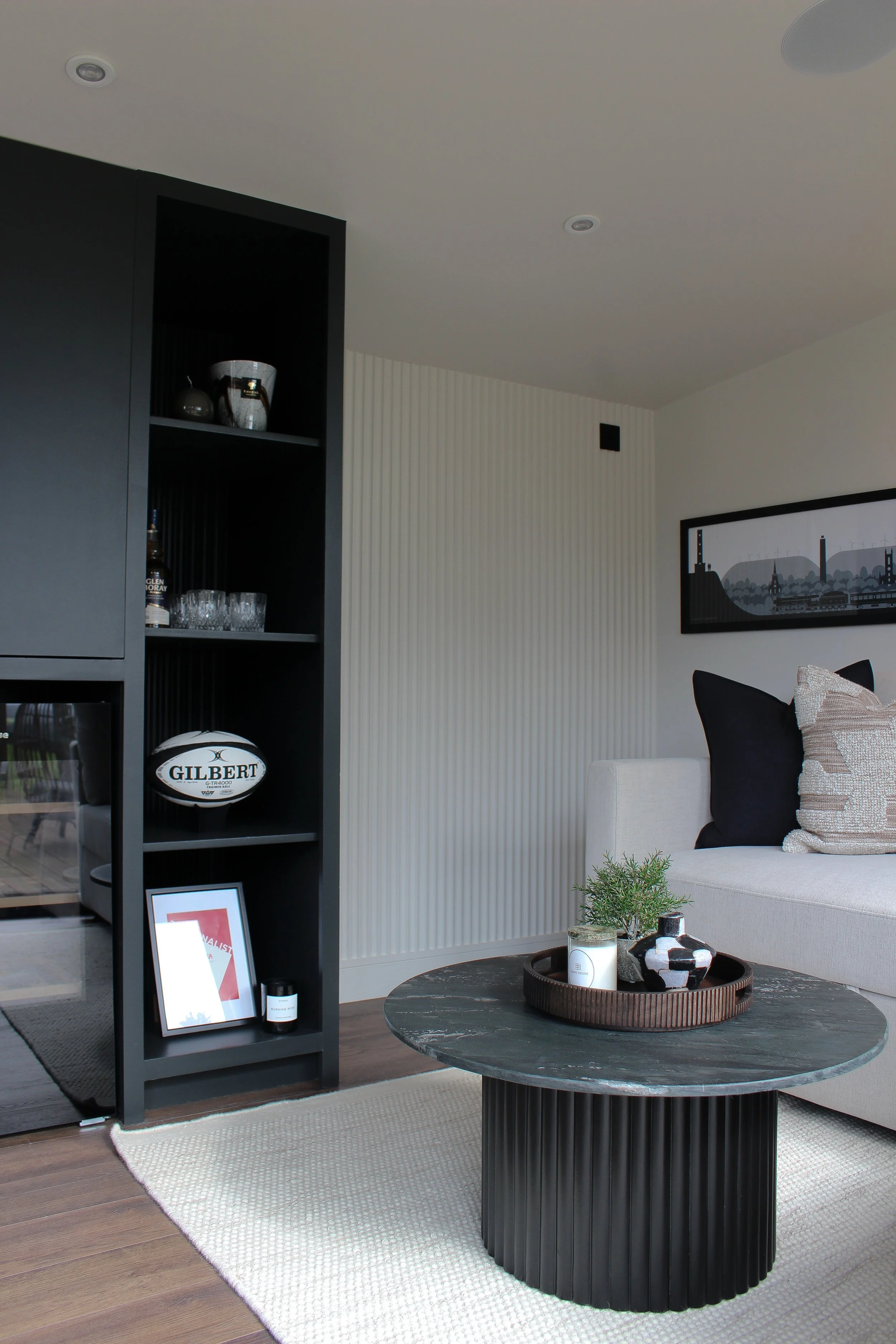 Office with a black bookshelf, white sofa with black and beige pillows, a round black coffee table with decorative plants and vases, framed artwork on the wall, and a beige wool rug on wood floor. Garden office designed by Yazzmin Lovelle Interiors.