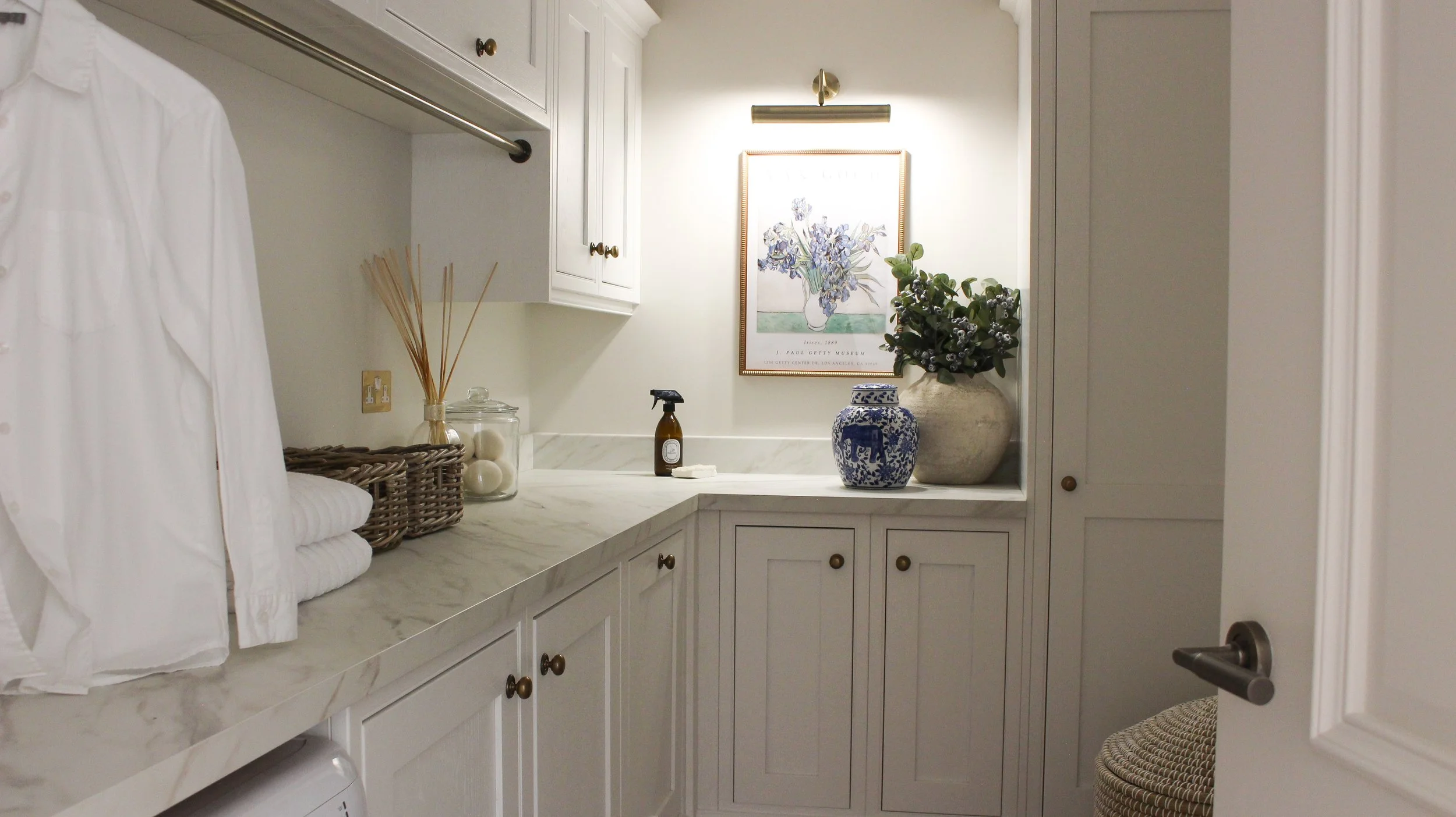 A laundry room with white cabinets, a marble countertop, and decor including vases, a framed floral art piece, and a potted plant.