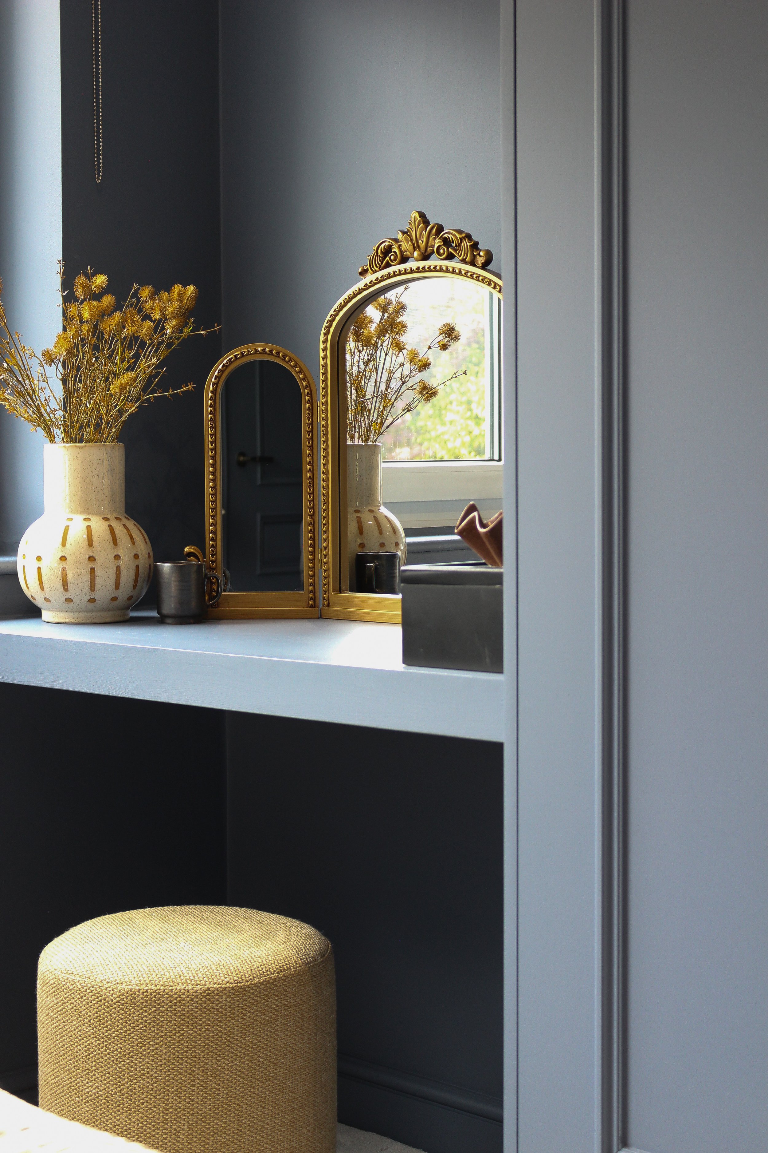 A vanity area with a gold ornate mirror featuring a beige vase with dried flowers, a small metallic cup, and a window in the background. There is a mustard upholstered stool below the countertop and dark blue cabinets surrounding the dressing table. 