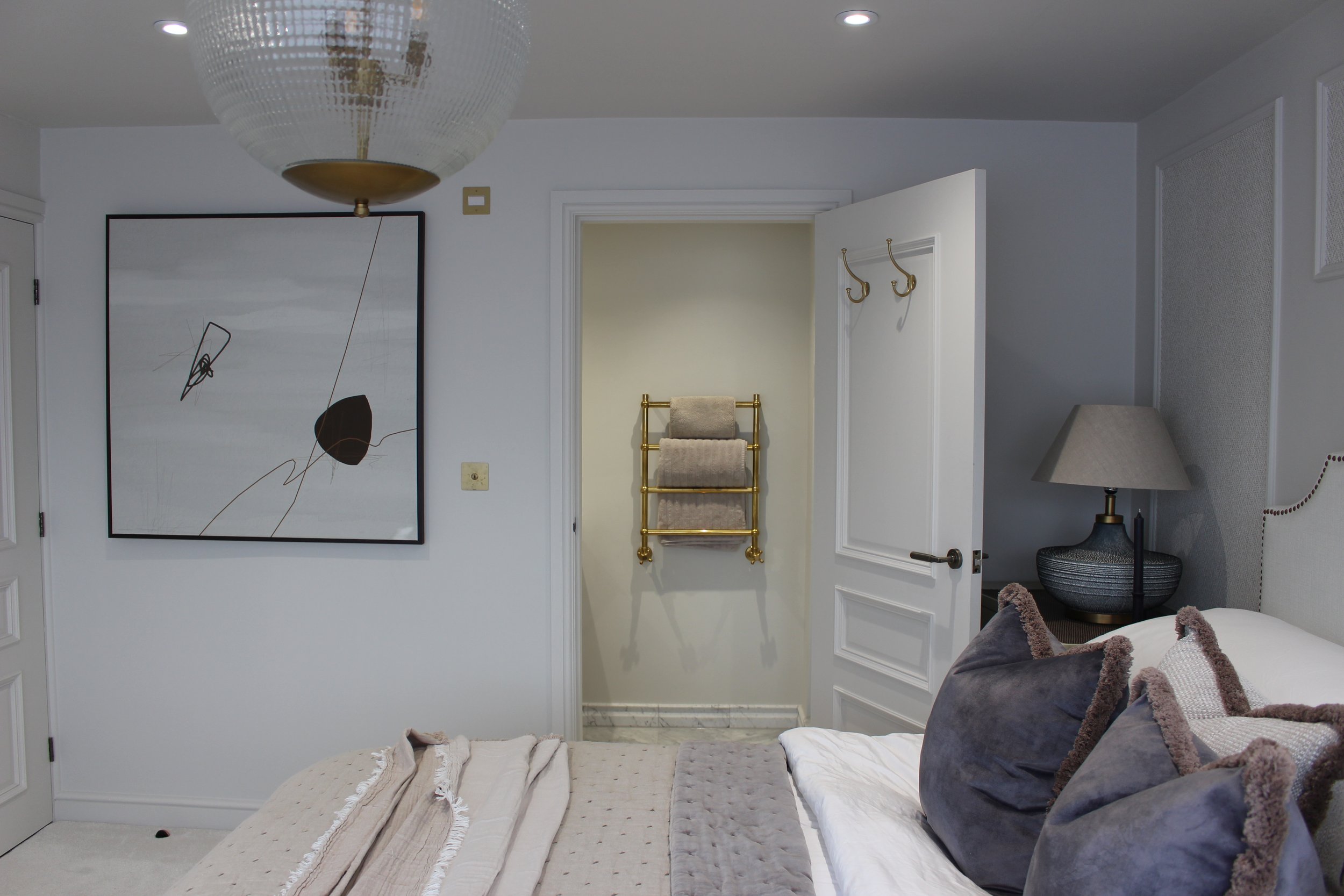 A bedroom with a bed, grey throw cushions, and a large bedside table with a lamp. An open door shows a small room ensuite with towels on a antique brass towel radiator. There is a large abstract painting on the wall and a ceiling glass light fixture.
