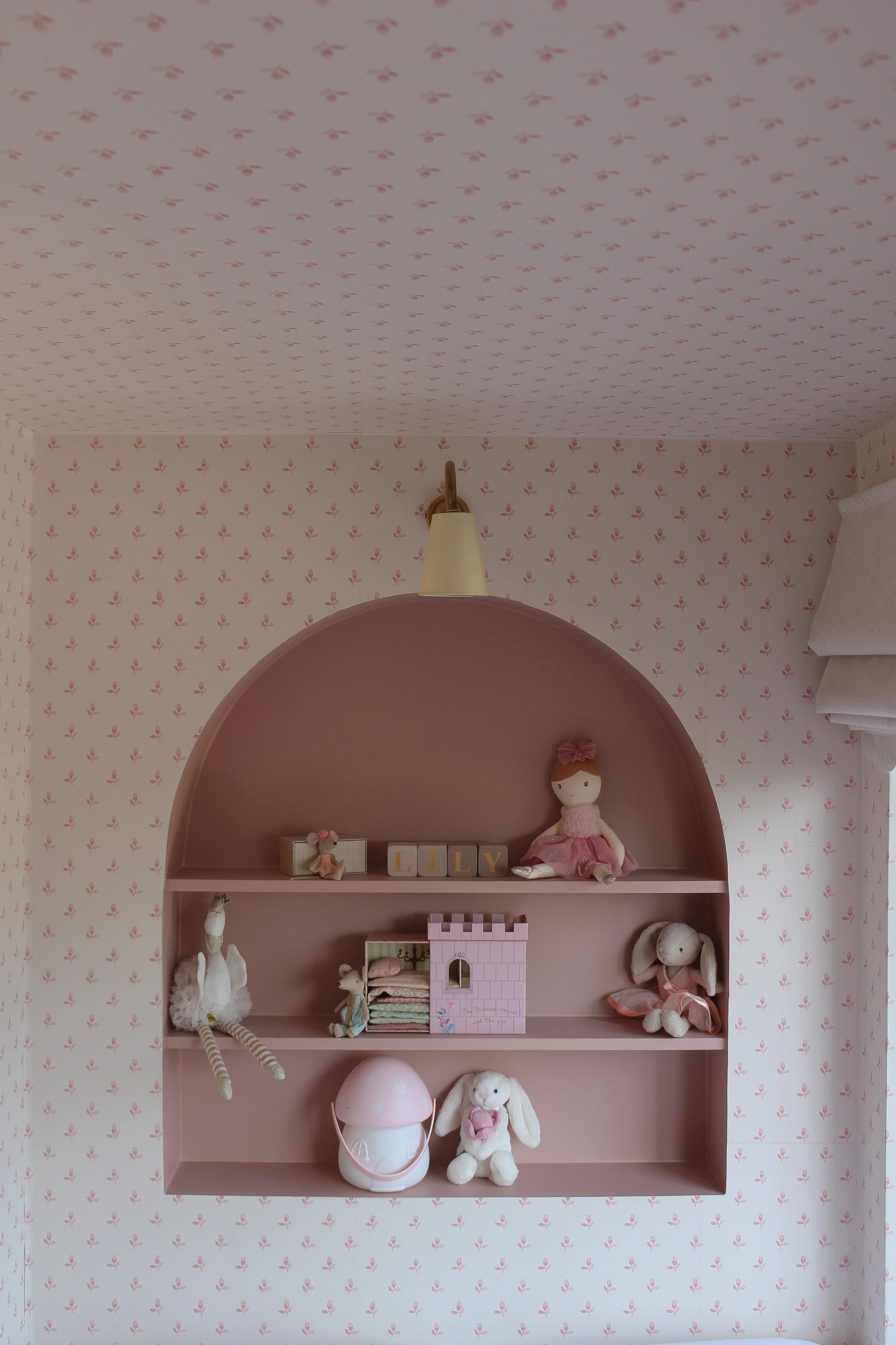 Pink girls bedroom wall with traditional print wallpaper and arched shelf with toys and decorations for a girl named Lily, including stuffed animals, dolls, and a mushroom-shaped lamp. Pink little girls room designed by Yazzmin Lovelle Interiors.