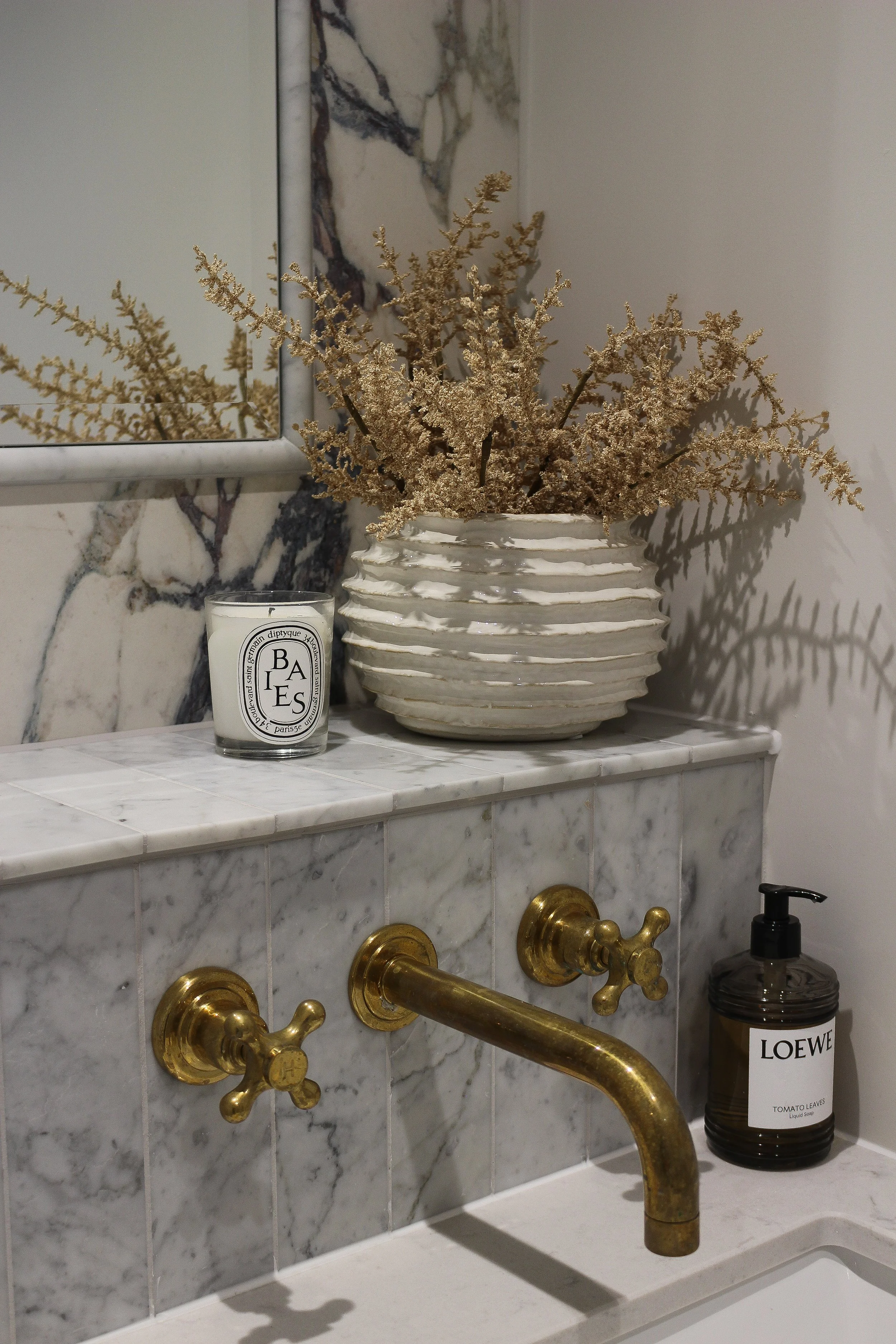 Bathroom sink area with marble countertop, gold tap and handles, a large decorative vase with dried branches, a branded Diptique candle, and a soap dispenser labeled 'LOEWE TOMATO LEAVES'.