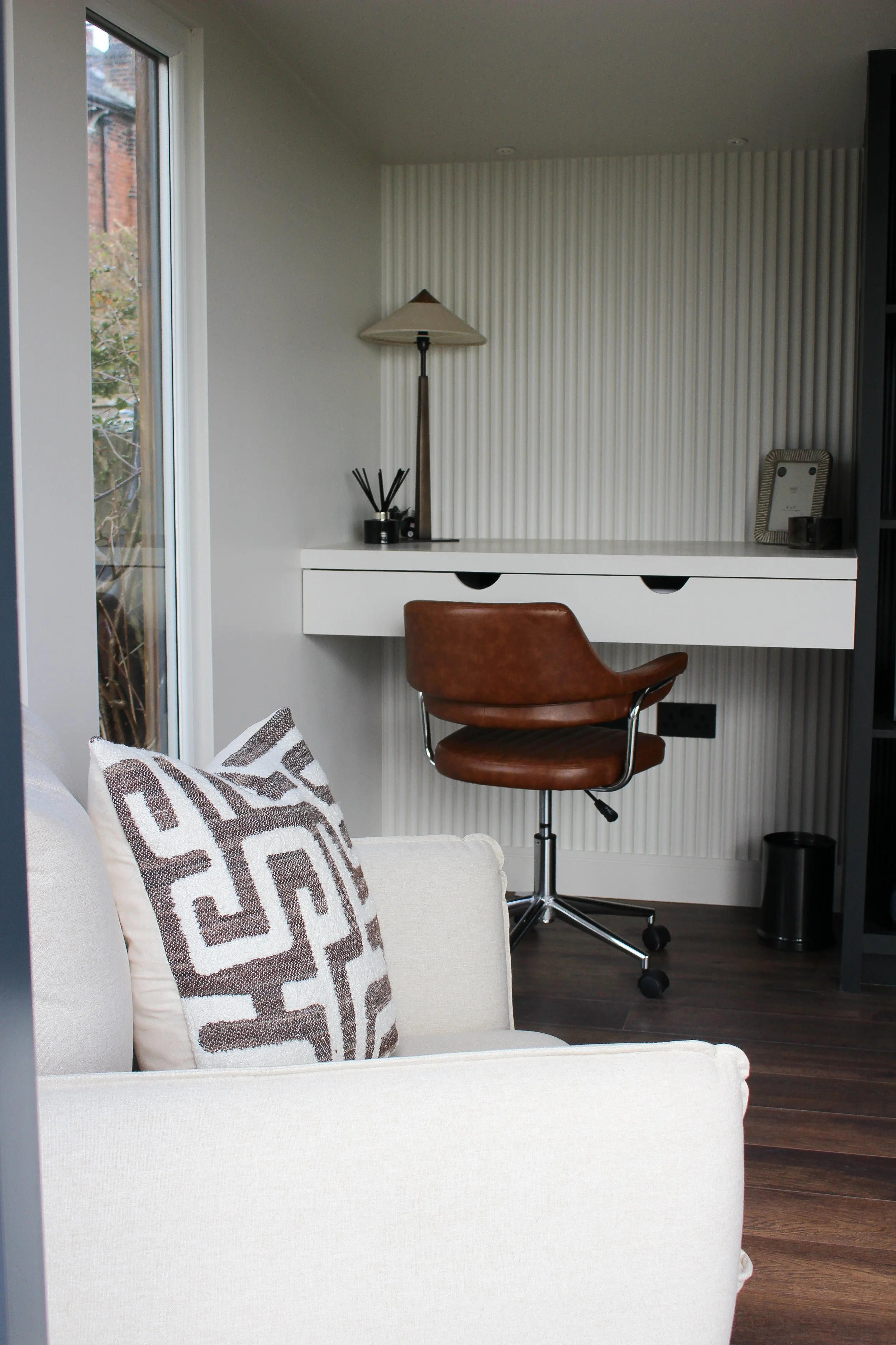 A corner of a office with a beige sofa and decorative pillow, a white desk with a brown leather office chair, a window, and a wall with textured white panels. Garden office designed by Yazzmin Lovelle Interiors.