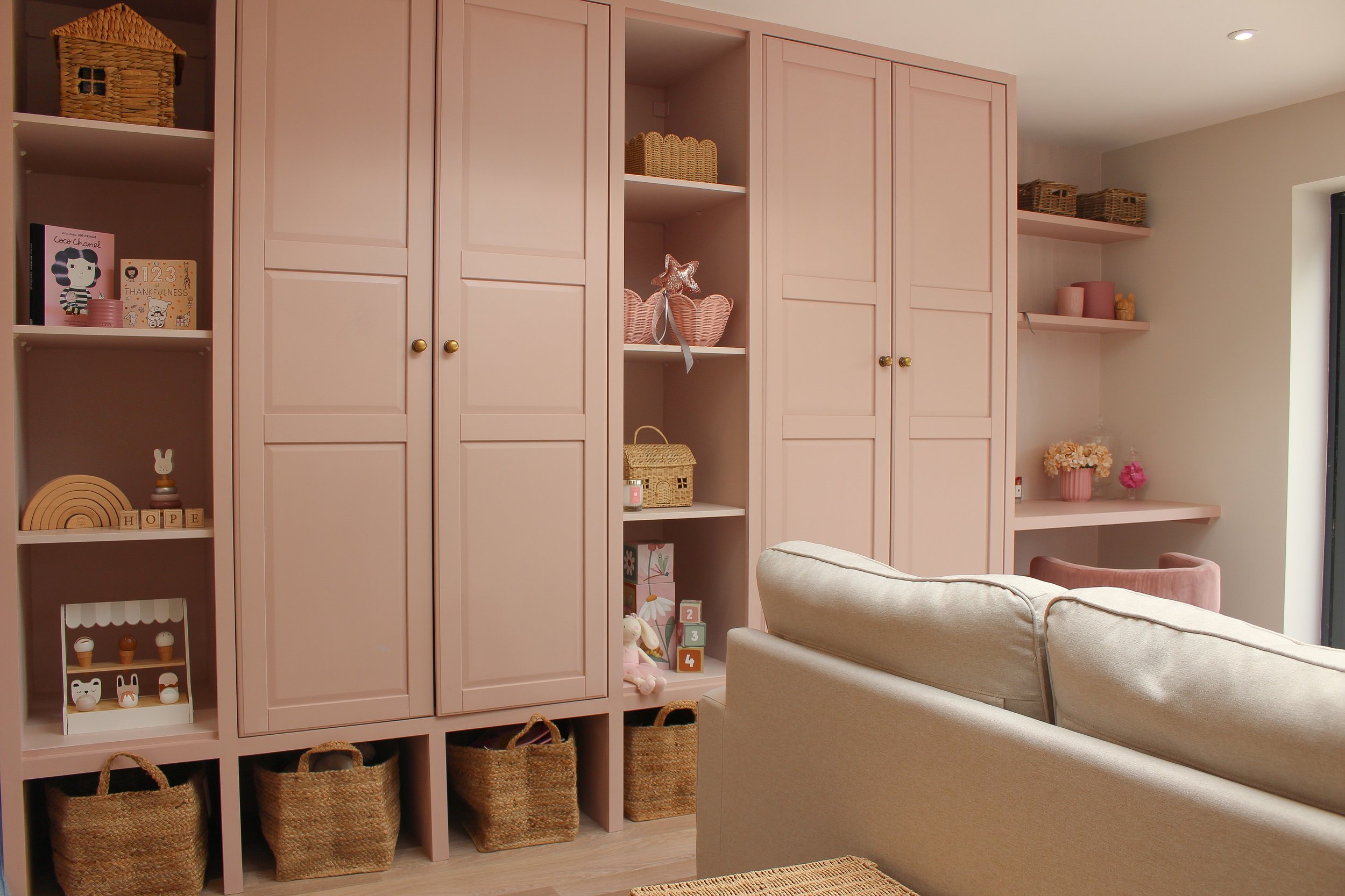 Pink built in closed storage with styled shelves, baskets, and decorative items, with a beige sofa in the foreground. Playroom designed by Yazzmin Lovelle Interiors.