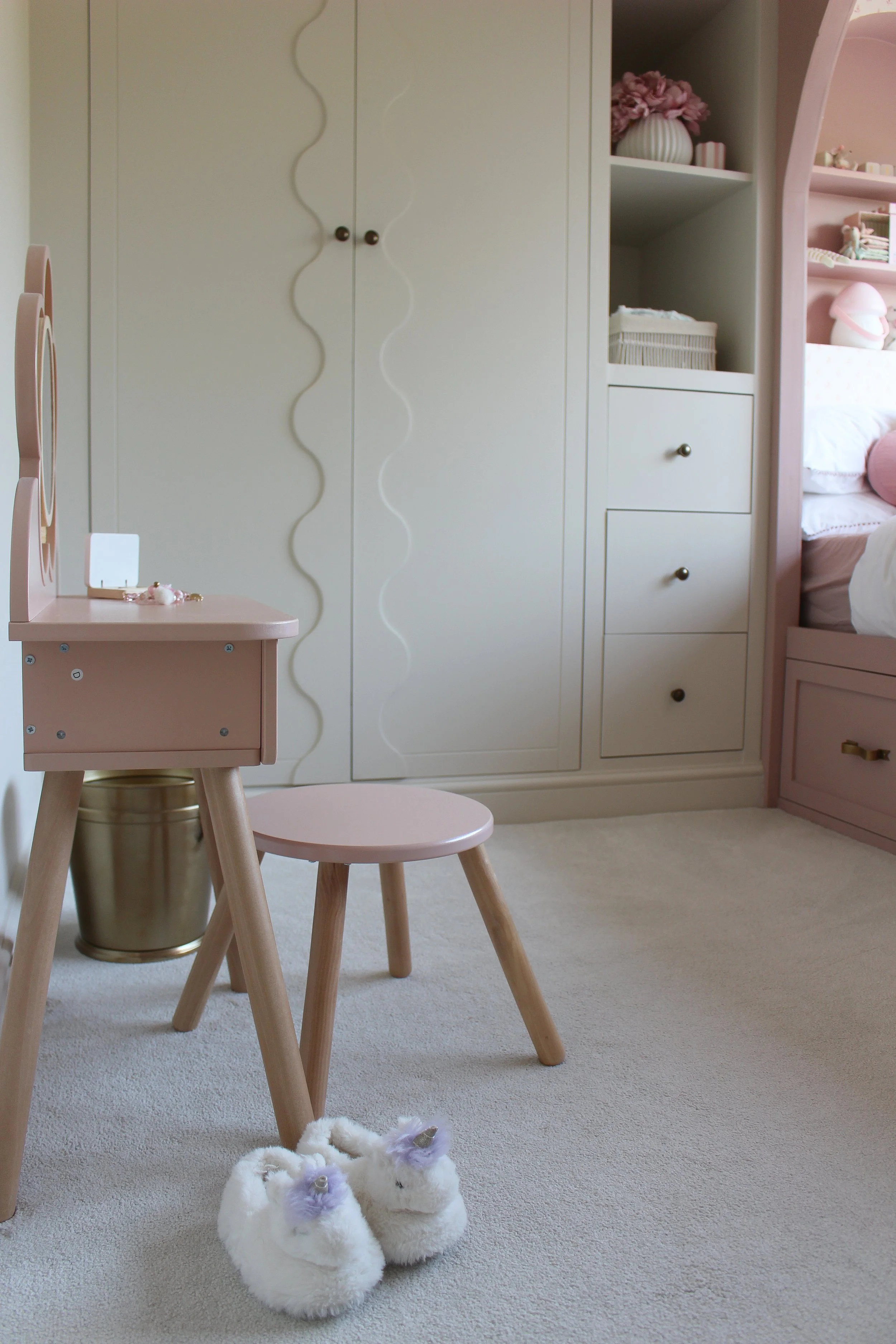 A cozy children's bedroom with pink and white decor, featuring a pink vanity, a matching pink stool, white plush unicorn slippers on a beige carpet, a white wardrobe with a wavy design, and a pink bed with decorative pillows. 