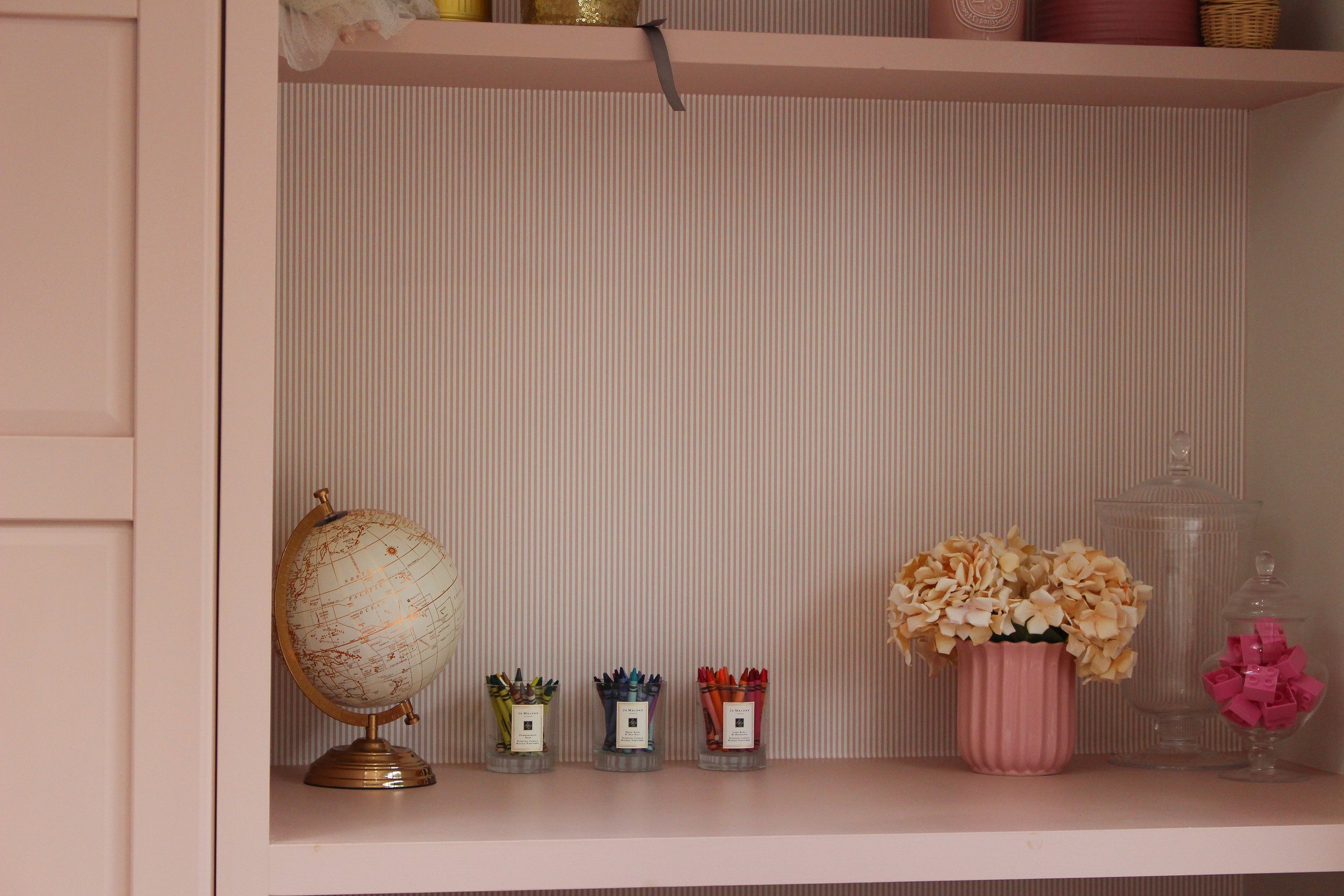 Decorative shelf with a miniature globe, three small containers of colored markers, a pink vase with cream coloured flowers, and two glass jars with lids, one filled with pink lego. Playroom designed by Yazzmin Lovelle Interiors. 