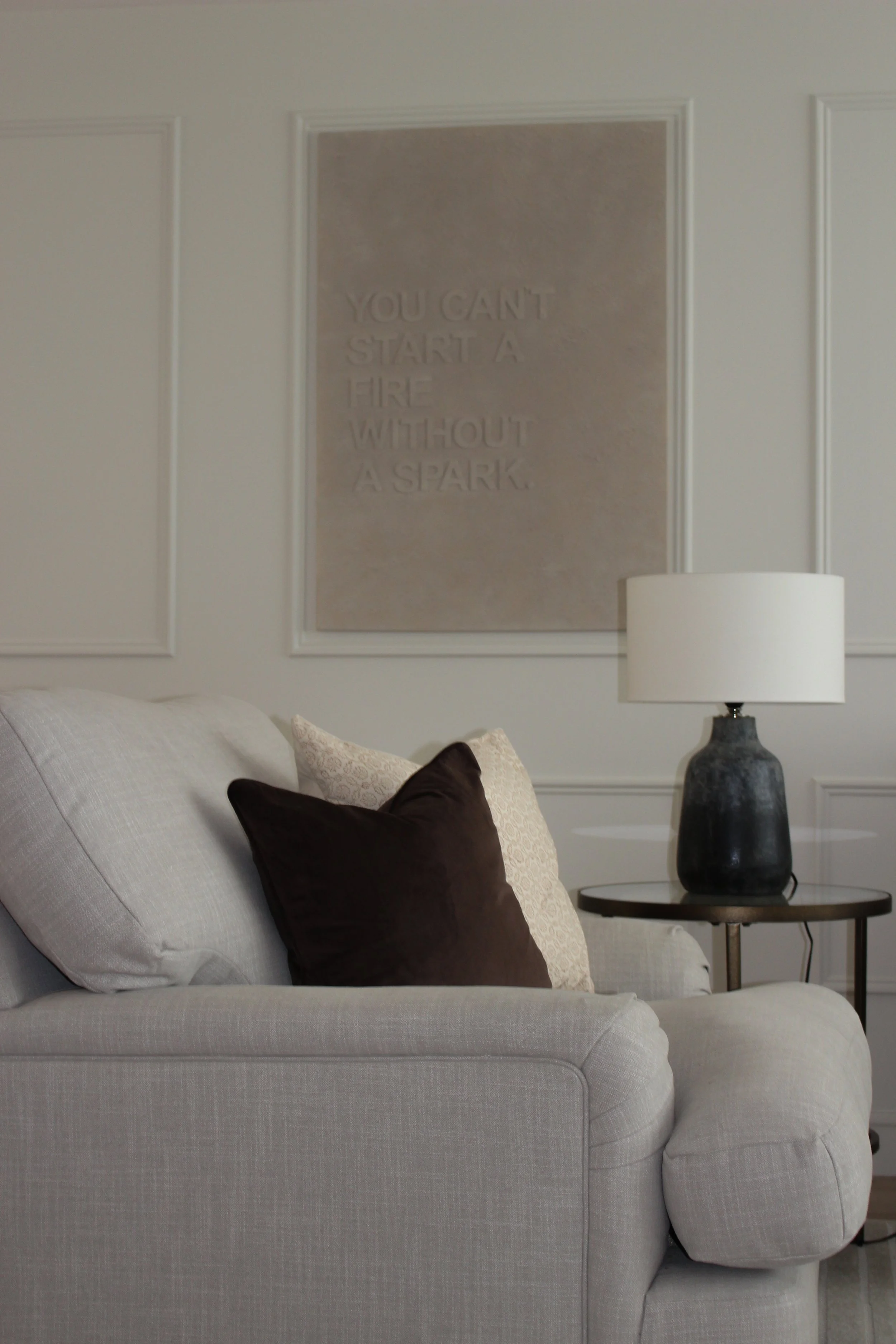 Living room with beige couch, three throw pillows, a black table lamp, and a wall art sign that reads 'You can't start a fire without a spark'.
