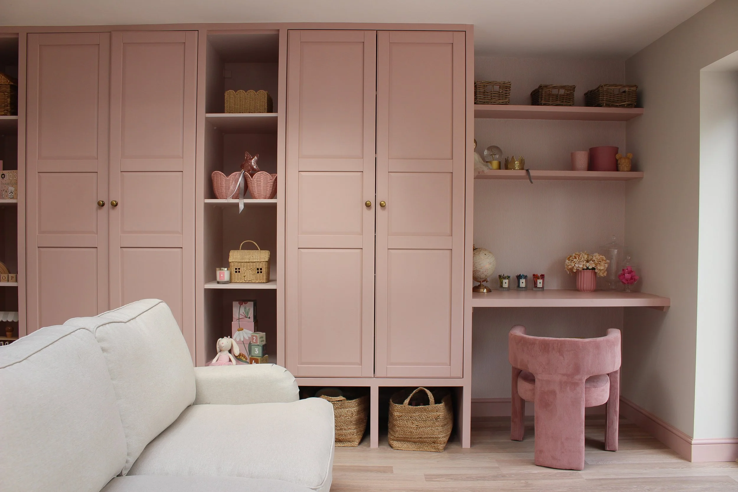 Interior view of light pink built-in playroom storage with shelves and closed cabinets, decorated with baskets, toys, and decorative items, alongside a pink velvet stool and a pink built in desk. Pink playroom designed by Yazzmin Lovelle Interiors.