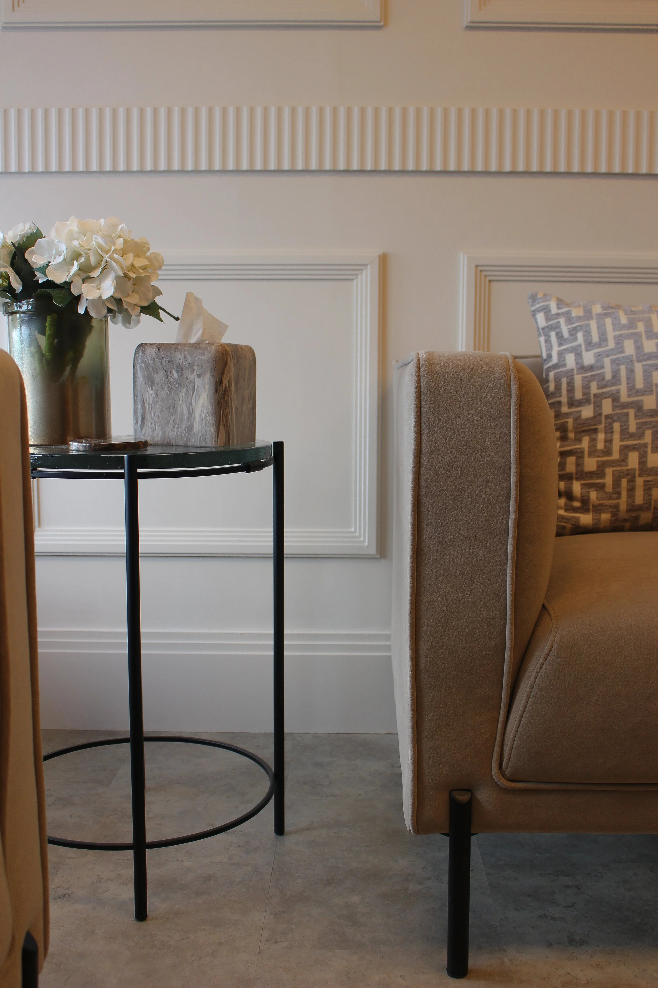 Close up of a beige arm chair with a decorative pillow, next to a black metal side table with a glass top holding a vase of white flowers and a stone tissue box, against a white panelled wall.