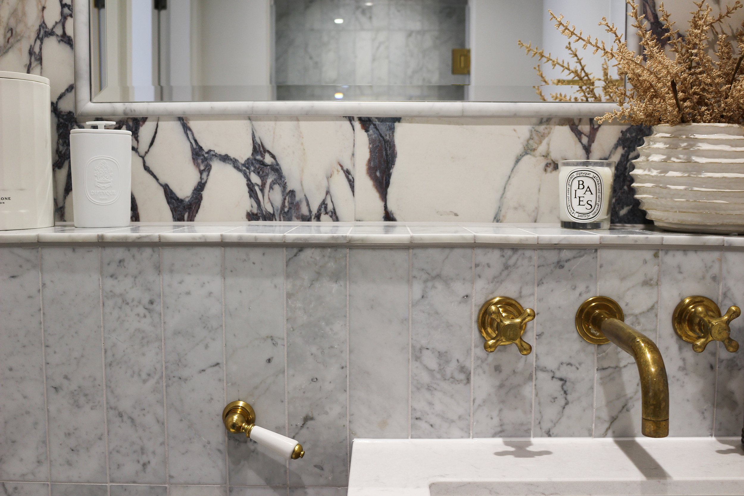 White marble bathroom with antique brass fixtures, a mirror, a soap dispenser, candle, and a decorative vase with dried flowers. Master ensuite designed by Yazzmin Lovelle Interiors