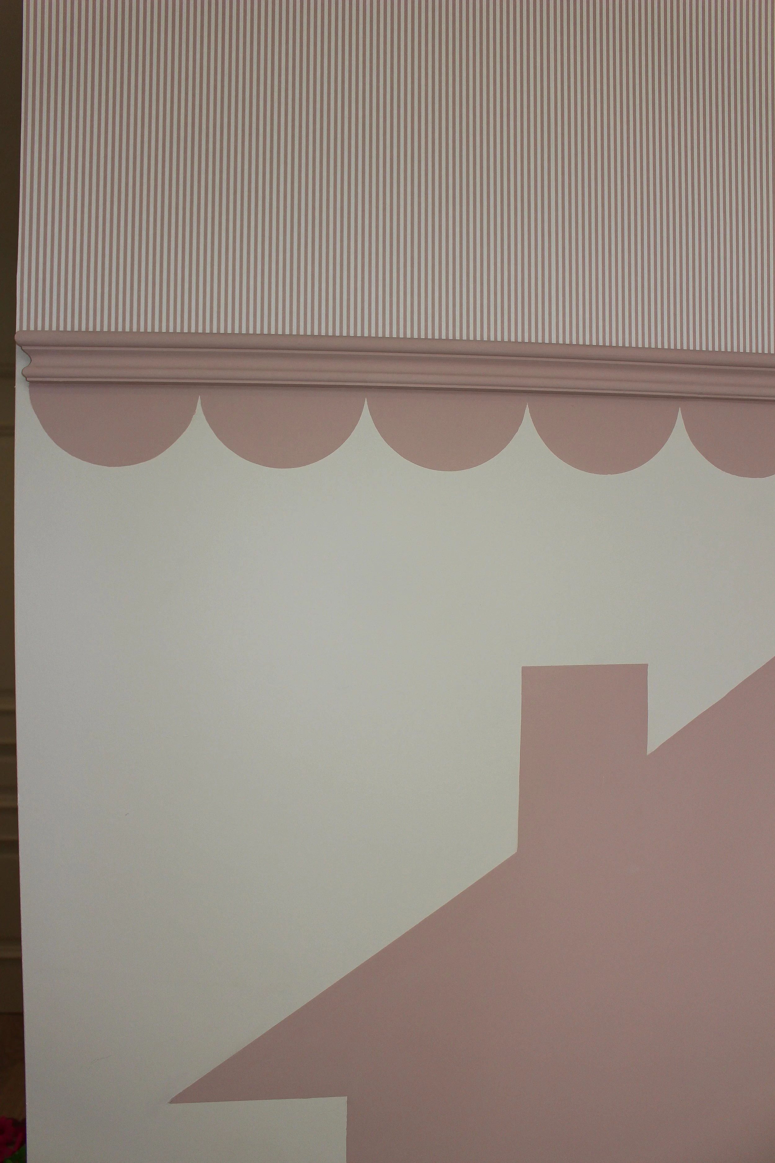 Close up of a wall with pink and white striped and scalloped design, including section of a pink house. Decorated with woodchip and magnolia paint and wallpaper. Playroom designed by Yazzmin Lovelle Interiors.