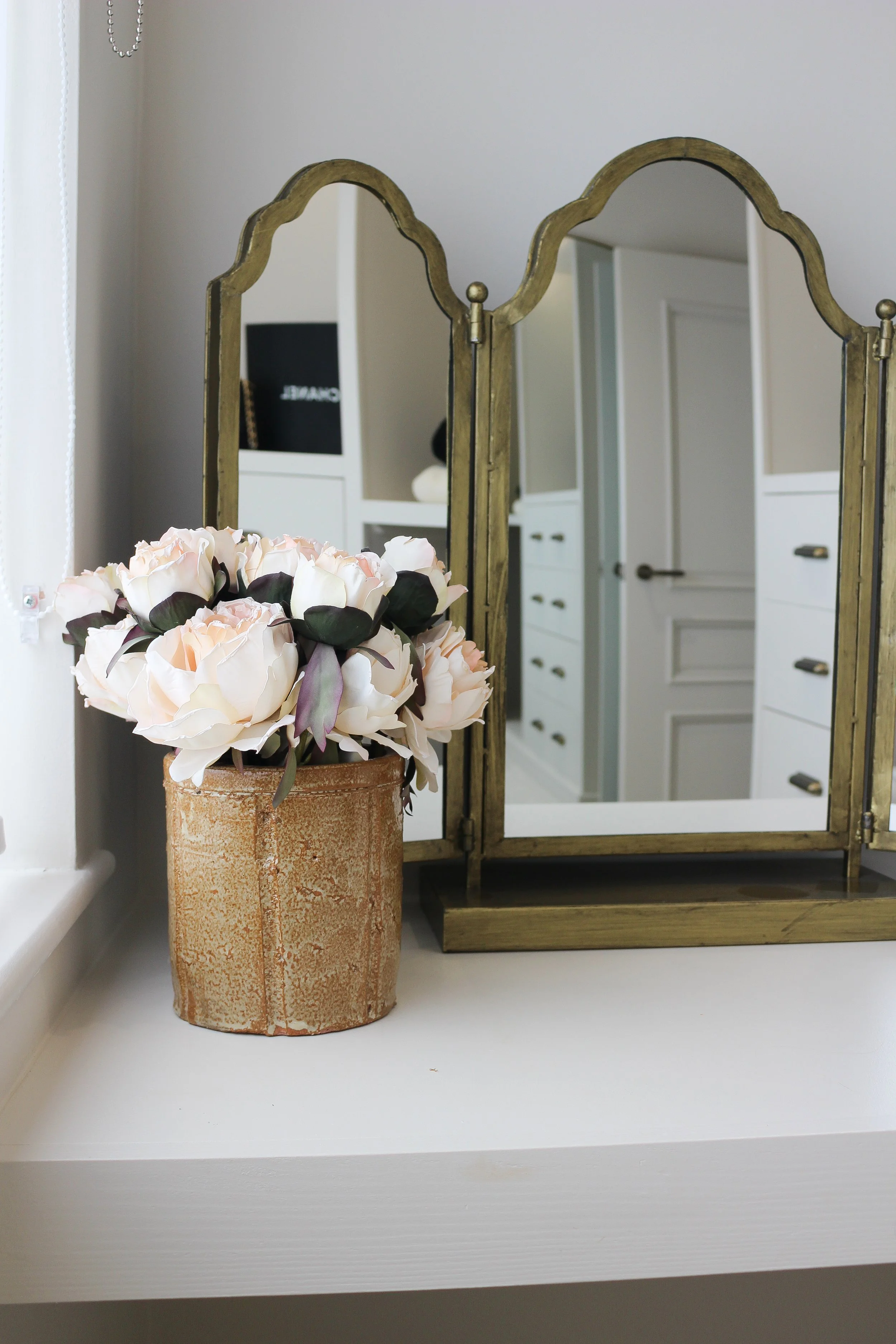 A decorative setup on a white surface featuring a rustic flower pot with pink and white peony flowers and a vintage gold folding mirror behind it, with part of a white cabinet and door visible in the background.
