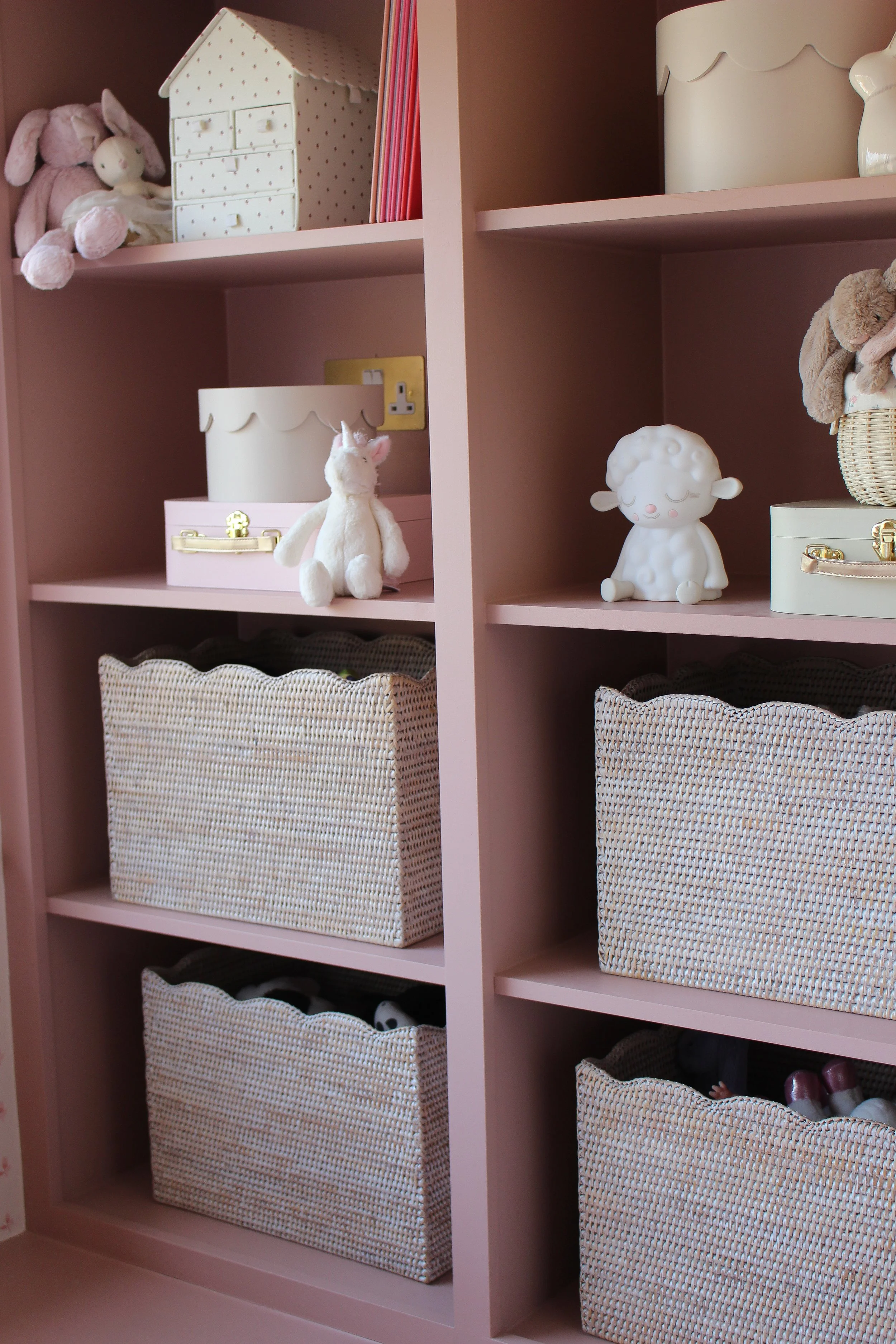 Pink shelving unit with woven baskets and plush toys, including a stuffed unicorn, a sheep figurine, and other stuffed animals. Girls bedroom designed by Yazzmin Lovelle Interiors