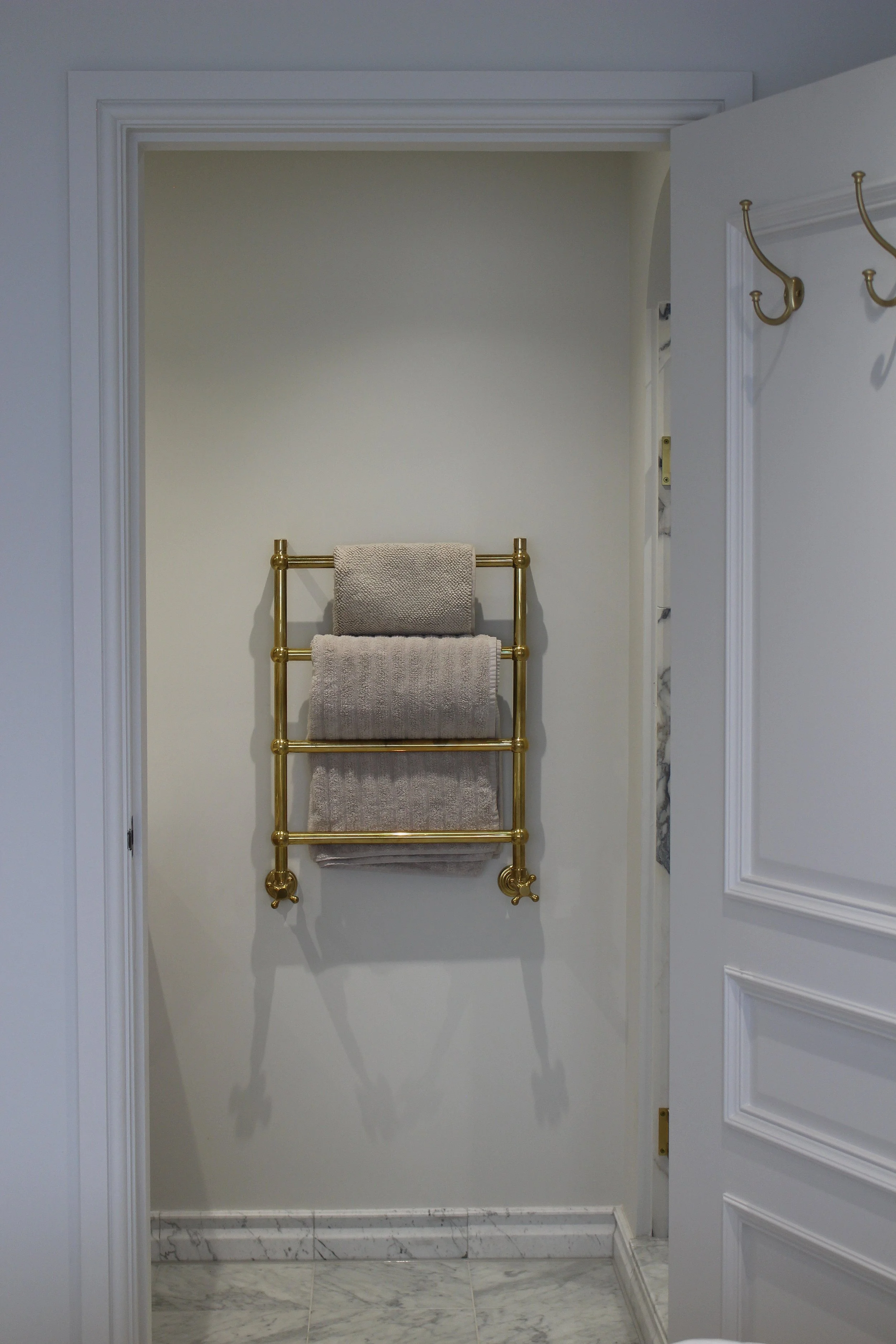 A antique brass towel radiator with four folded towels hanging inside a ensuite bathroom, seen through a partly open door with decorative moulding, on a marble floor. Master bedroom designed by Yazzmin Lovelle Interiors